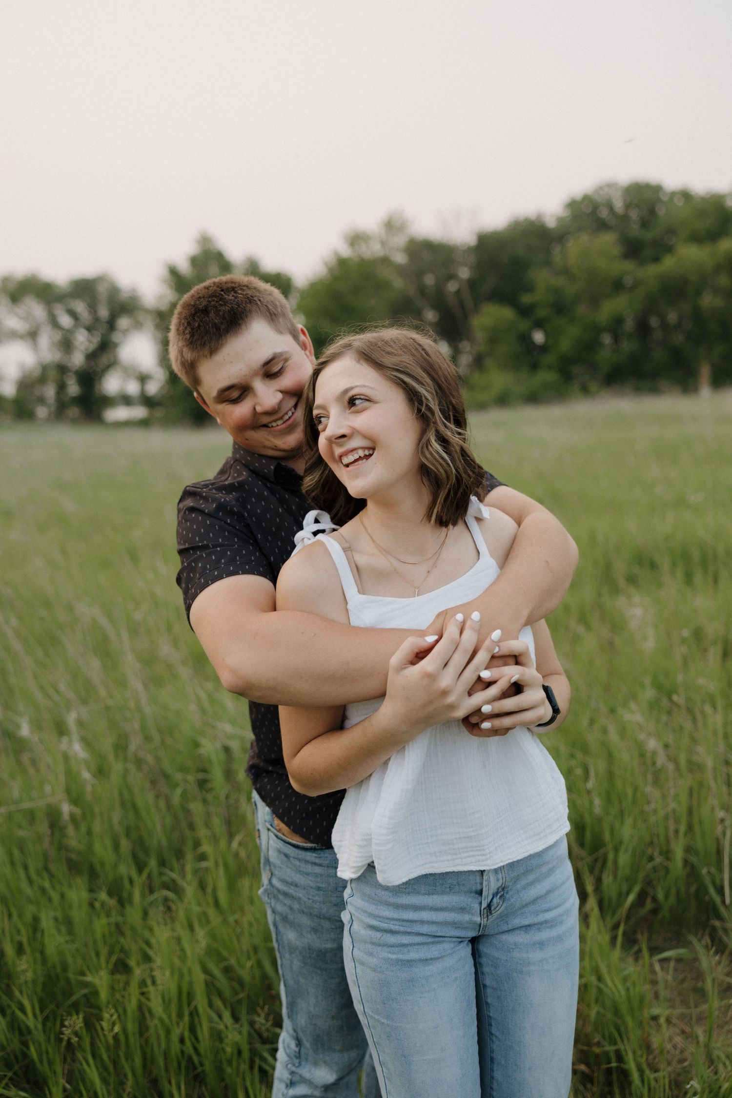 Joyful couple embracing and laughing together in a green meadow, woman in white top and jeans smiling up.