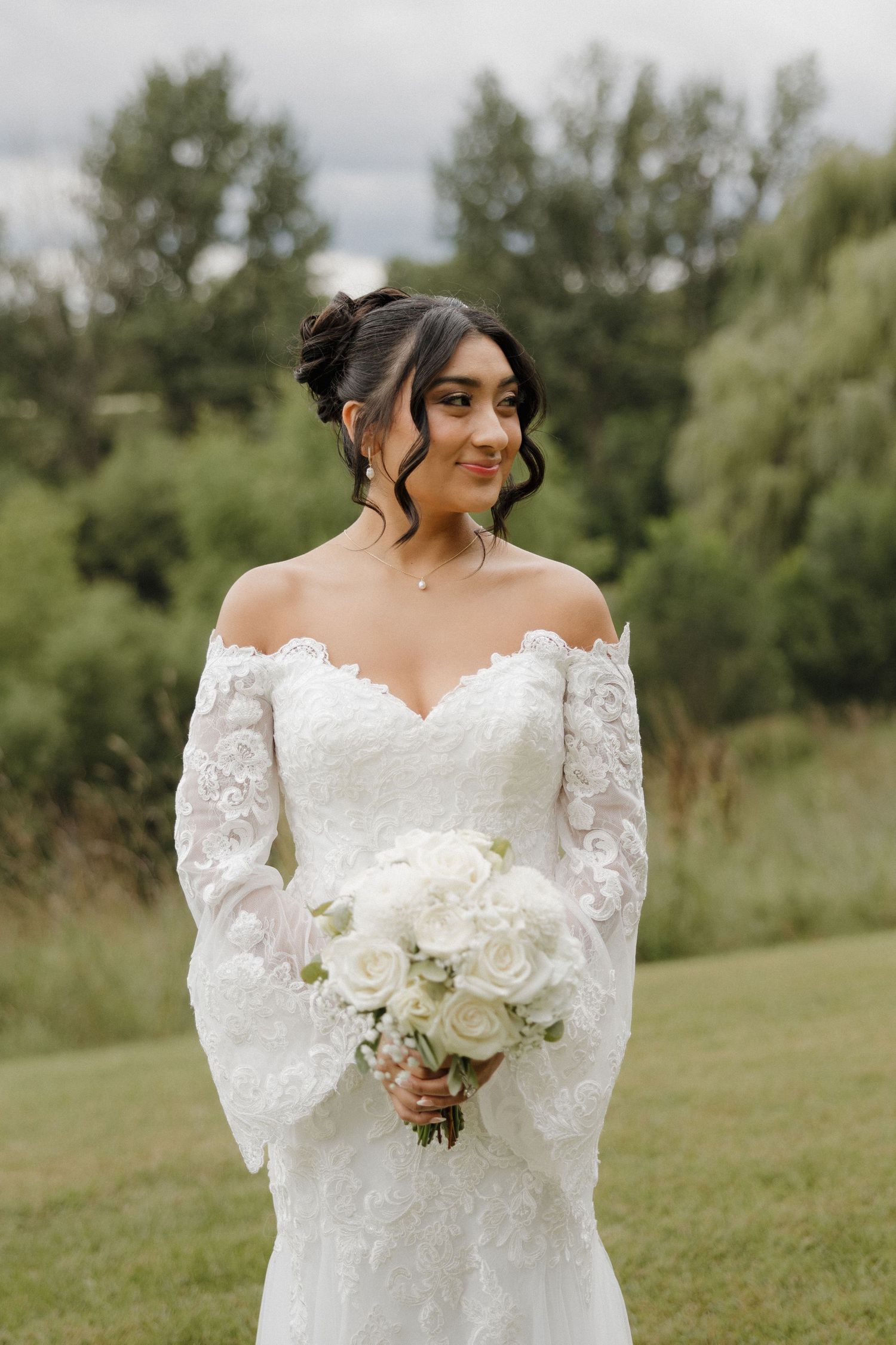 Elegant bridal portrait outdoors featuring off-shoulder lace wedding dress and white rose bouquet.