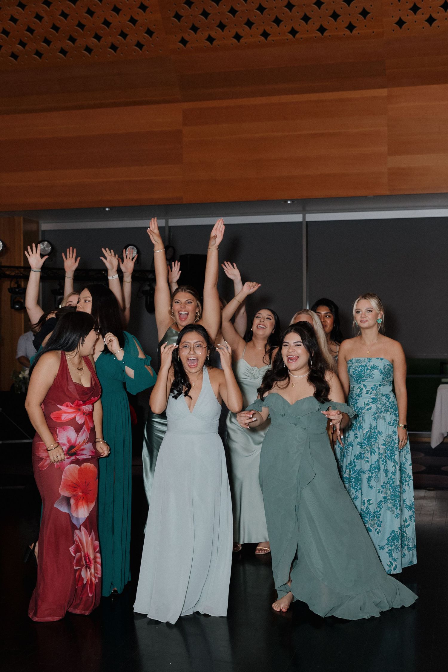 Group of wedding guests in colorful dresses dancing with raised arms at reception.