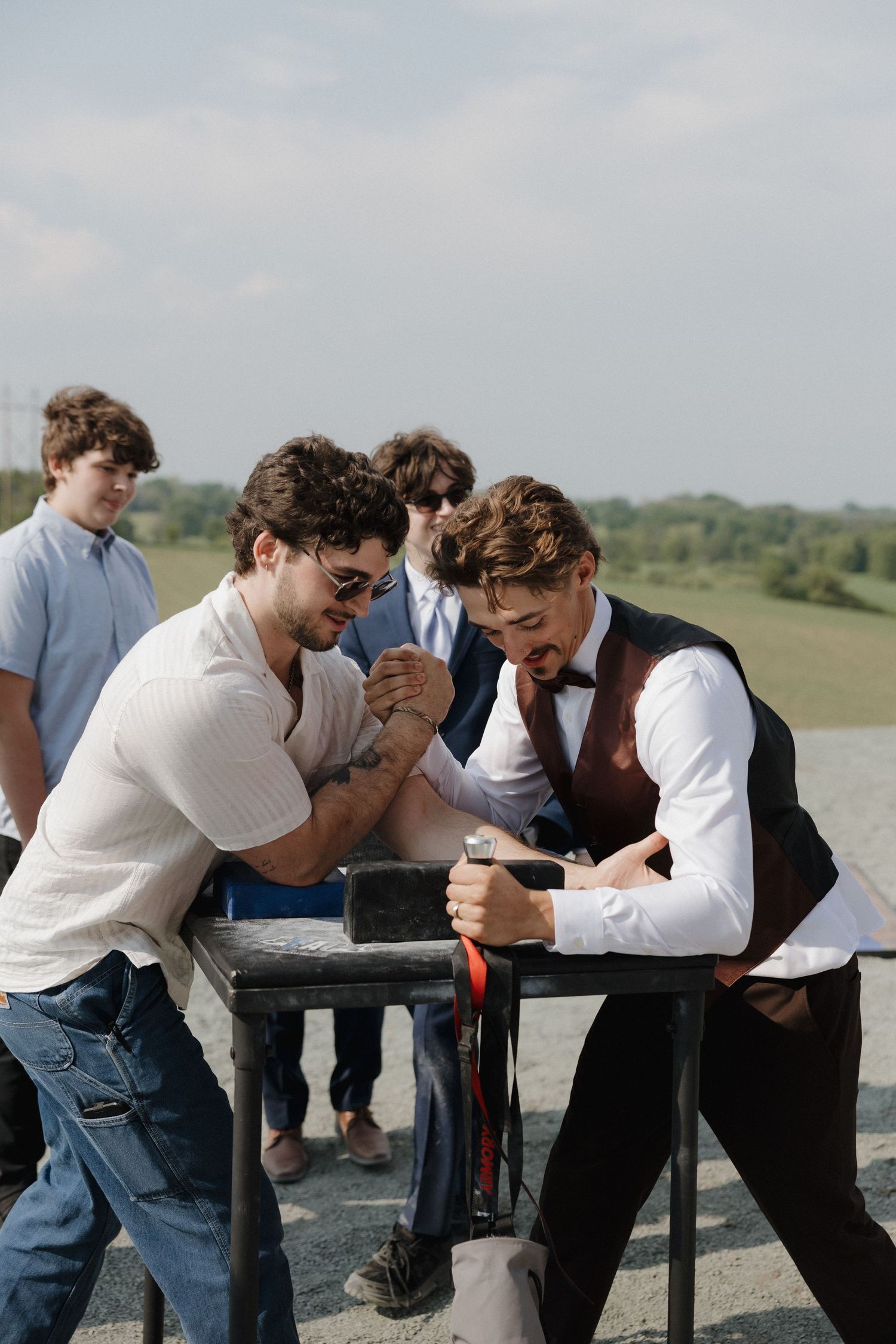Group of groomsmen in casual attire playfully arm wrestling outdoors.