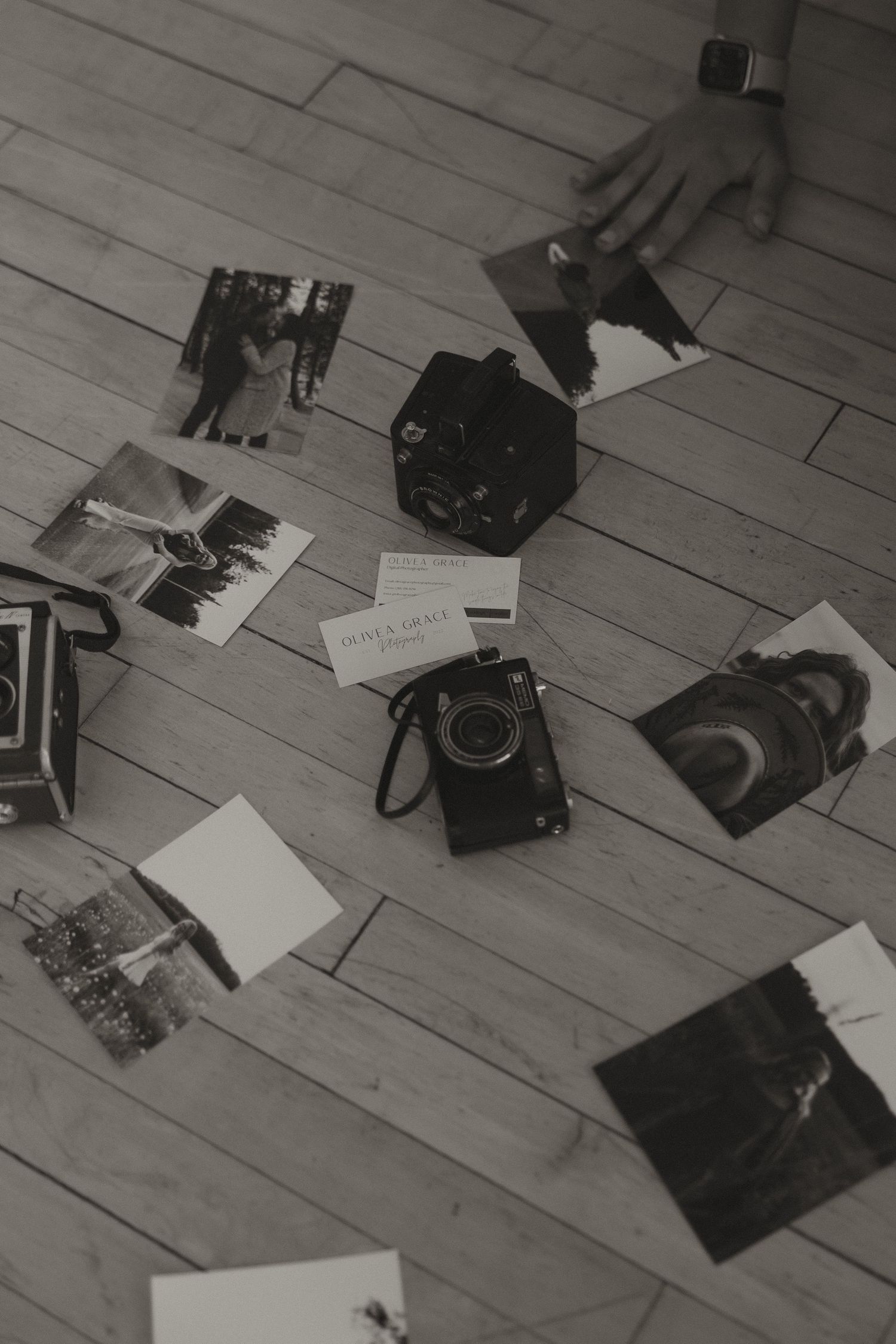 Black and white overhead photo of vintage cameras, printed photographs, and business cards on wooden floor.