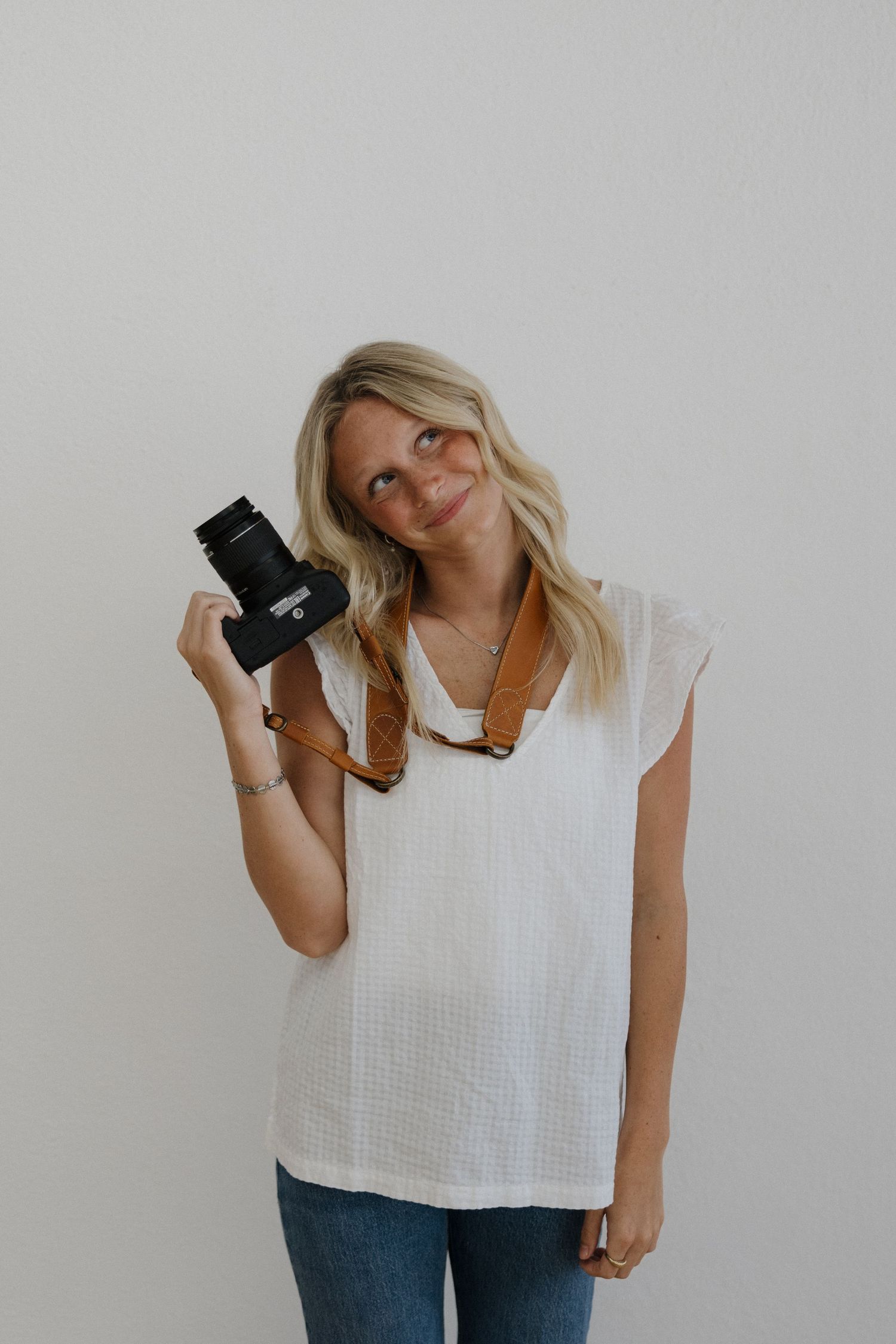 Blonde woman in white top holding a DSLR camera up while smiling in a bright studio.