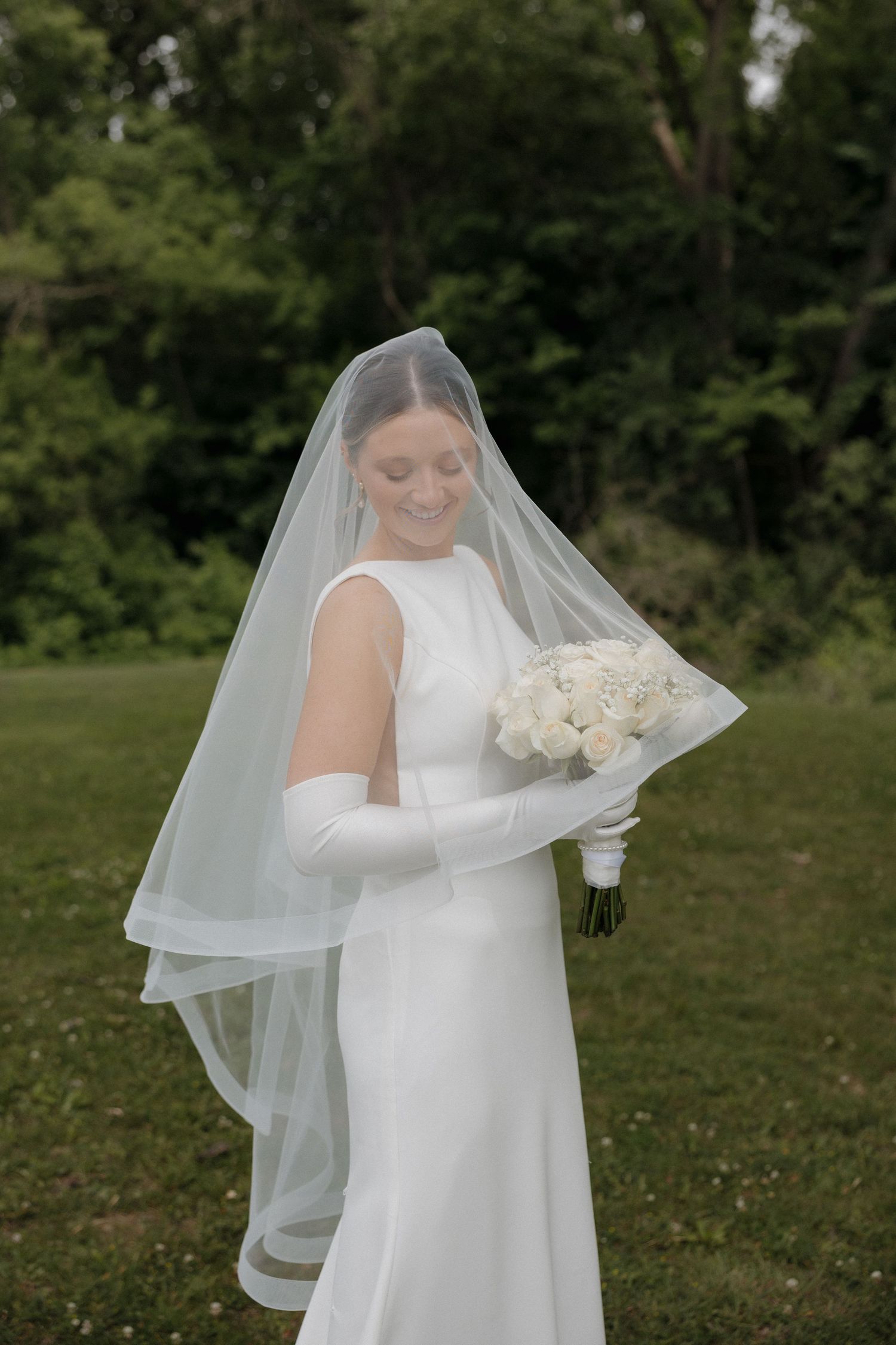 Bride in white sleeveless gown, long veil, and gloves holds white rose bouquet while smiling outdoors.