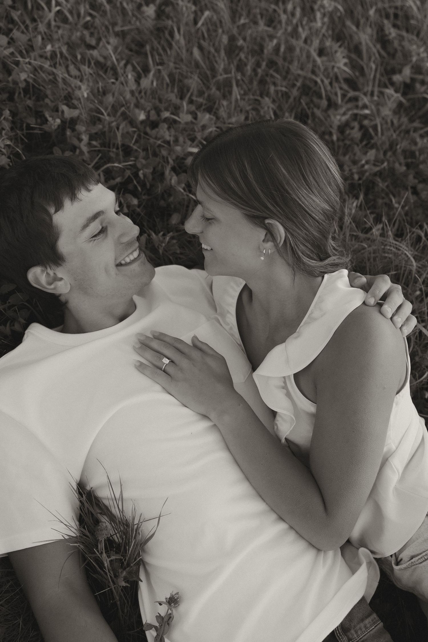 Black and white photo of a smiling couple lying in grass, gazing at each other lovingly.