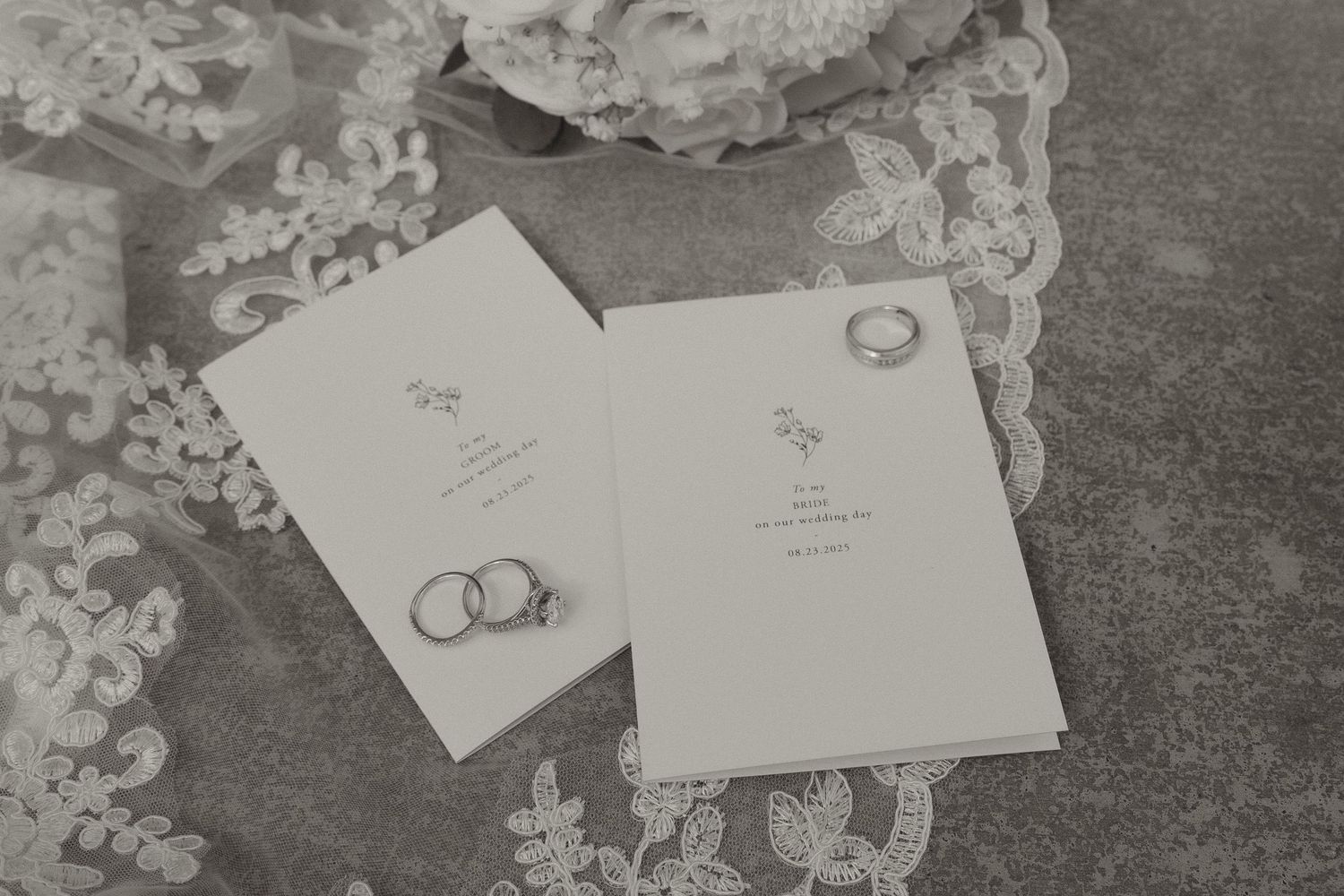 Wedding invitation cards with rings displayed on delicate lace fabric in black and white.