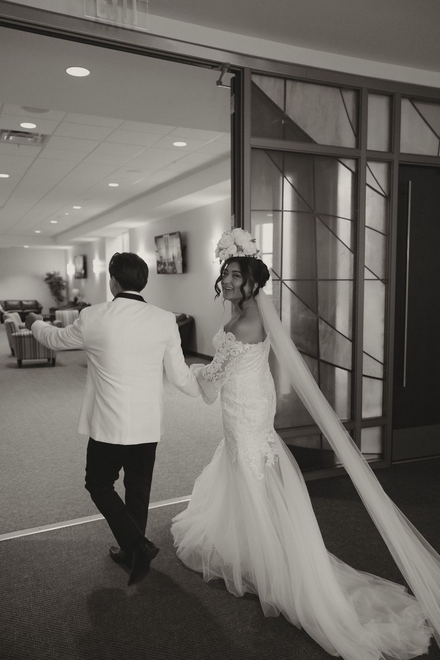 A black and white photo of a couple walking down a hallway in wedding attire.