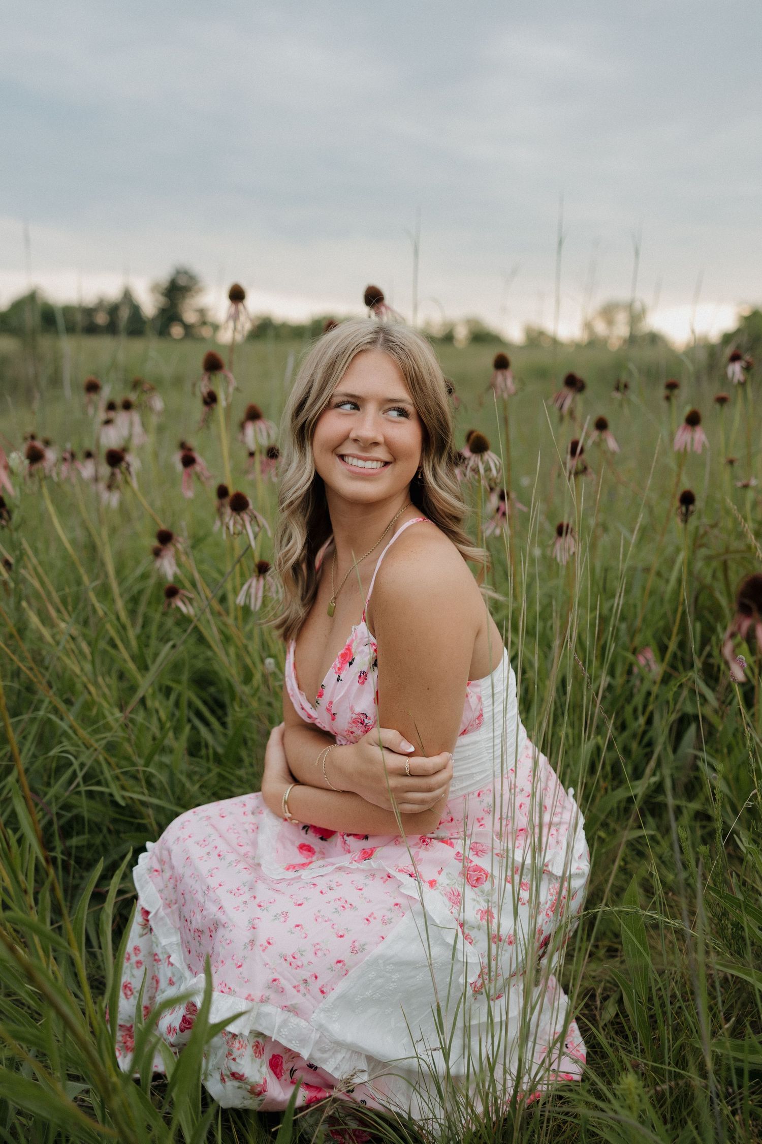 Smiling blonde woman in pink floral dress crouches in wildflower meadow on cloudy day.
