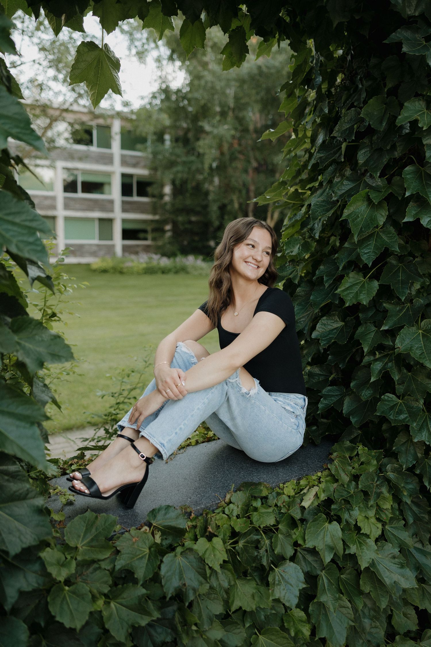 Young woman in black top and light jeans sits smiling among lush green ivy leaves outdoors.