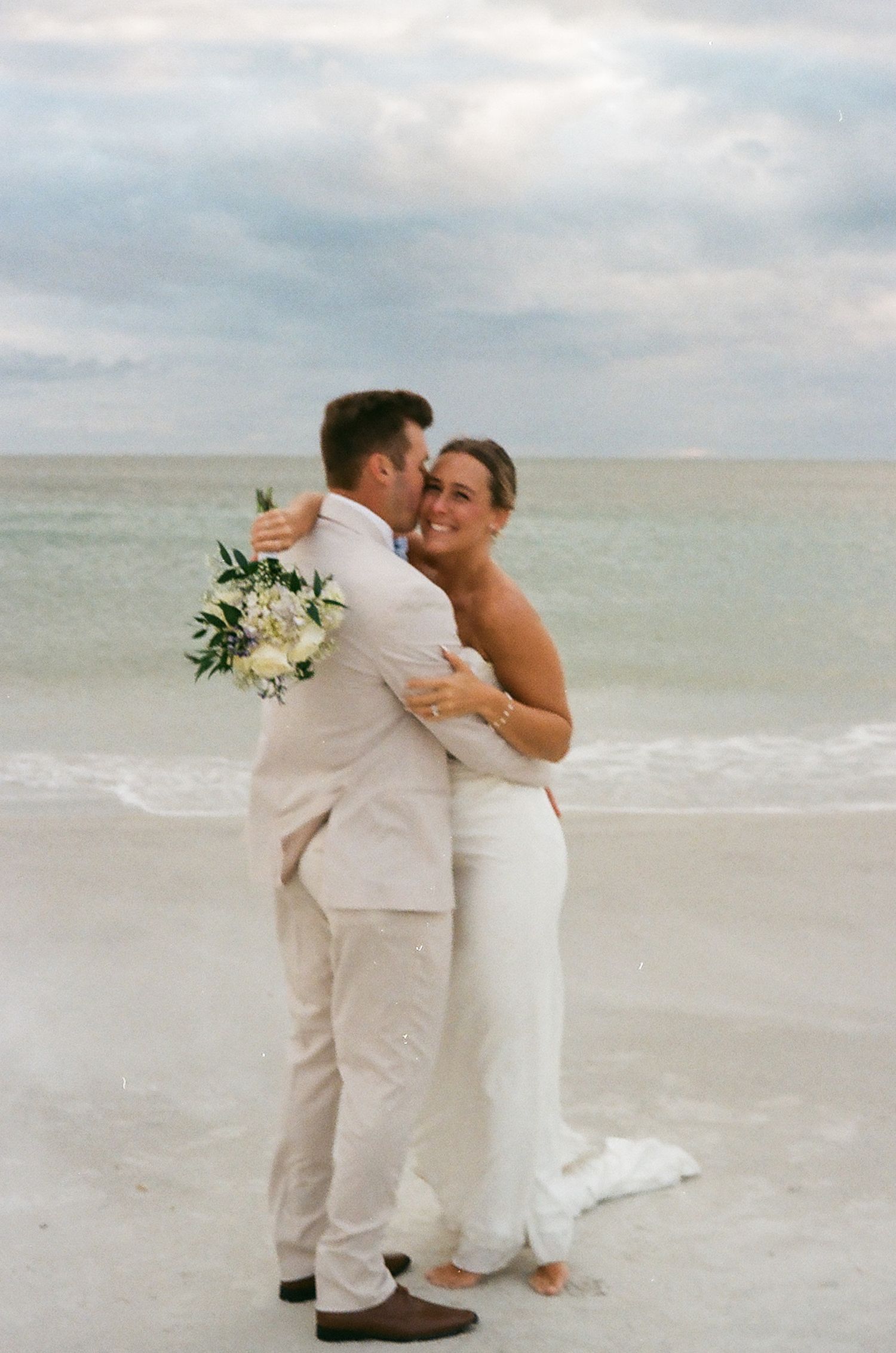 Bride and groom embrace on a sandy beach, she holds white bouquet, both dressed in white wedding attire.