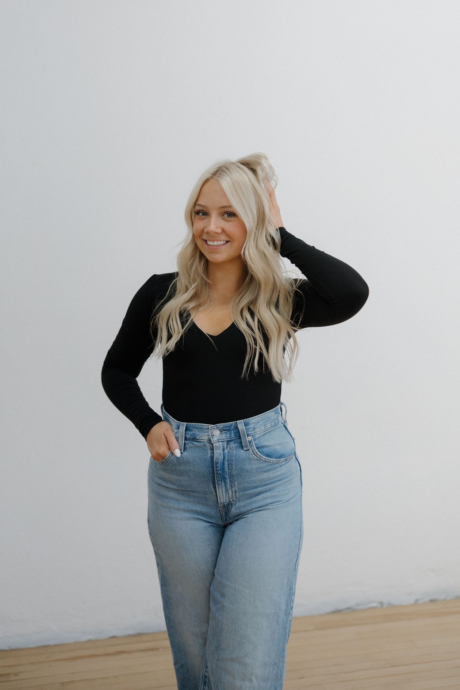 Smiling blonde woman in black bodysuit and high-waist jeans posing against white wall.