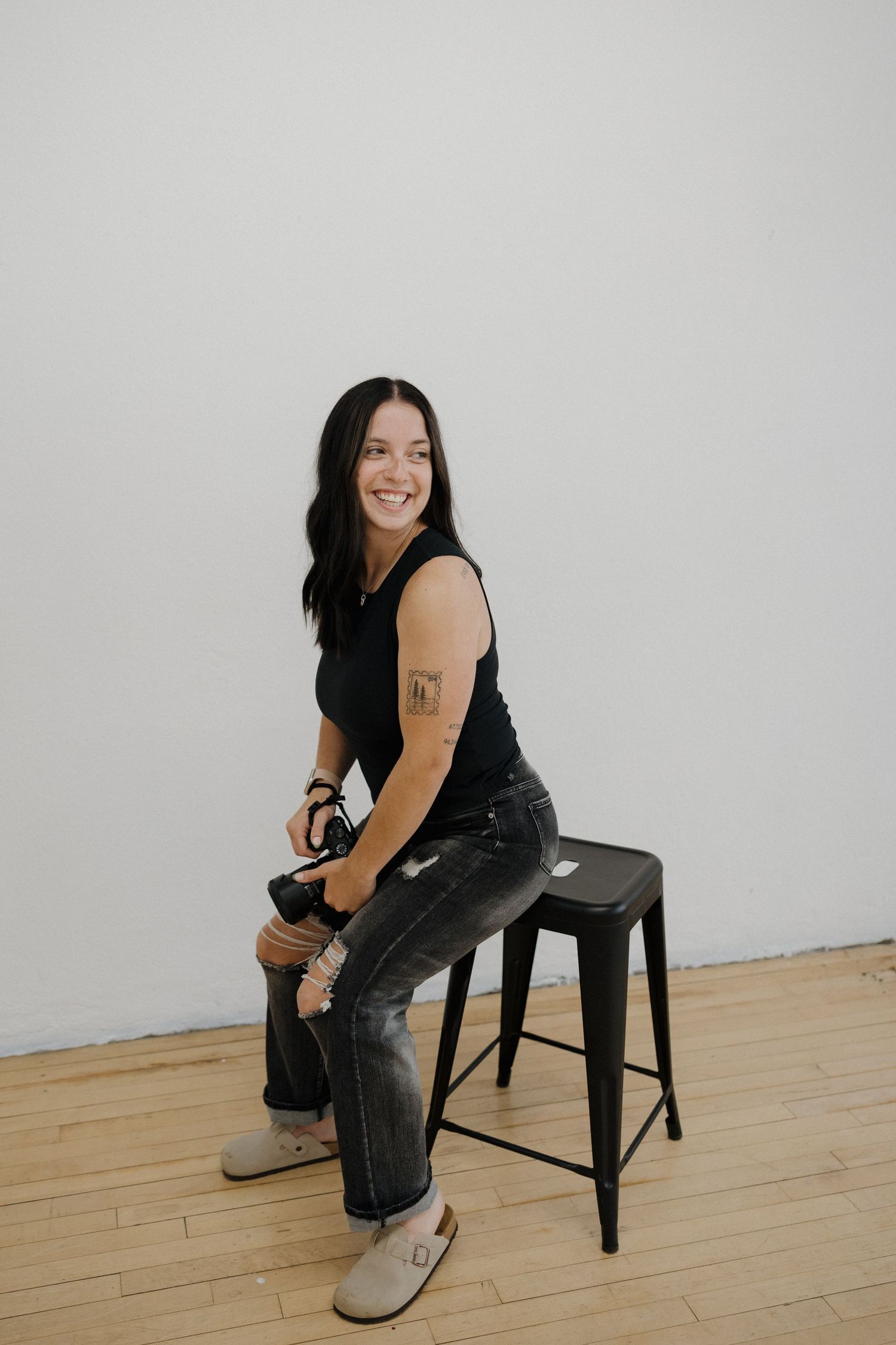 Smiling brunette woman sitting on a metal stool holding a camera in a bright studio.