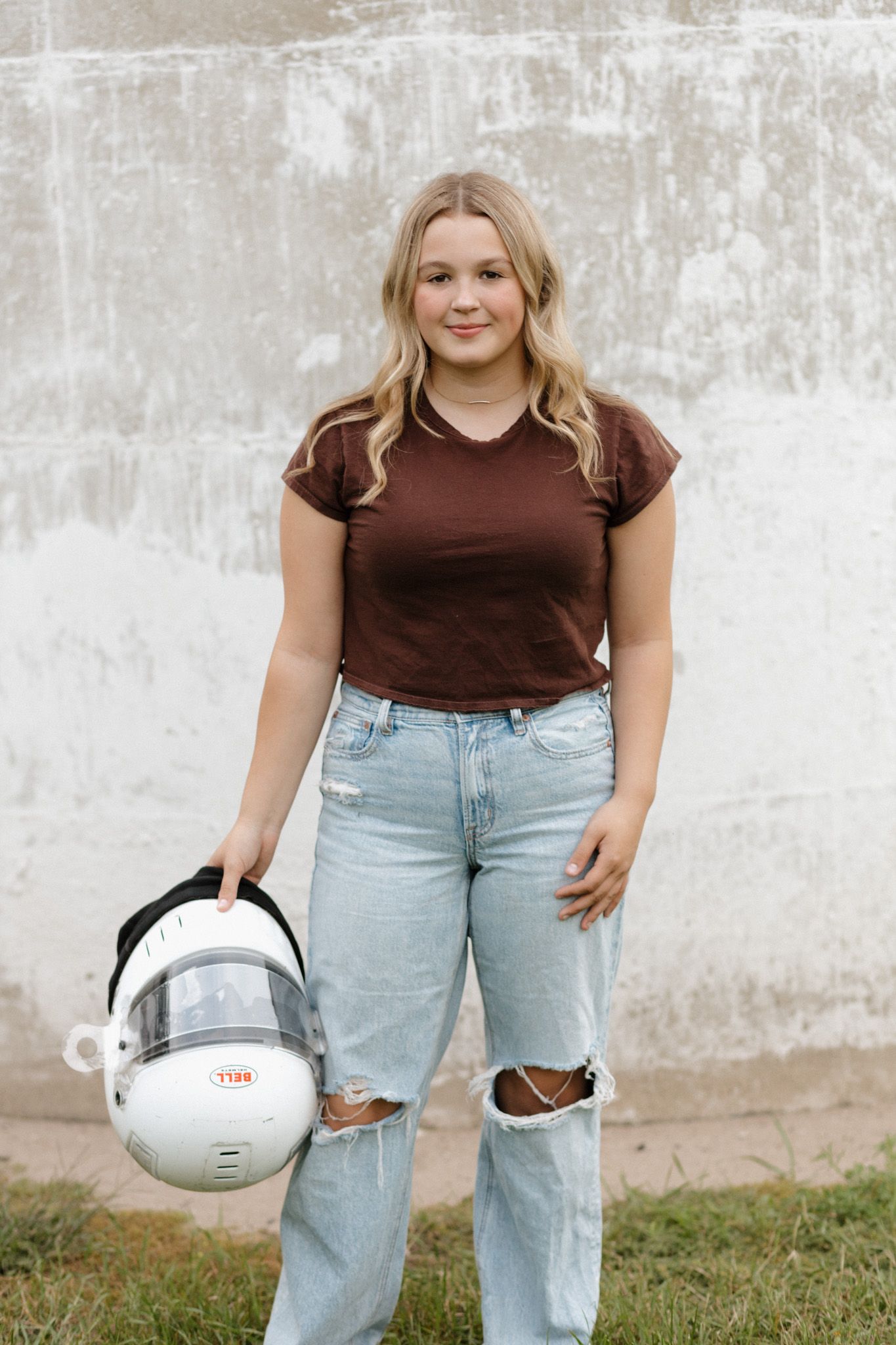 Blonde teen girl in brown tee and ripped jeans holding white racing helmet against concrete wall.