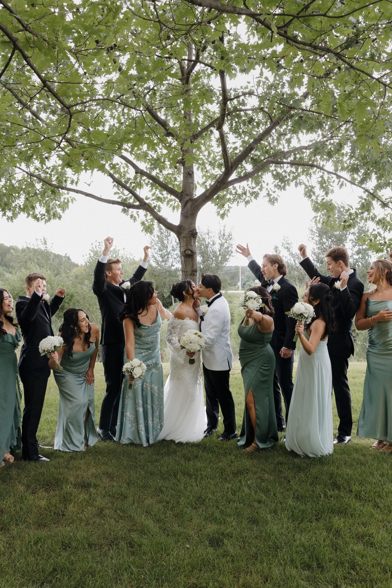 Large wedding party lined up outdoors under trees taking fun group photos together.