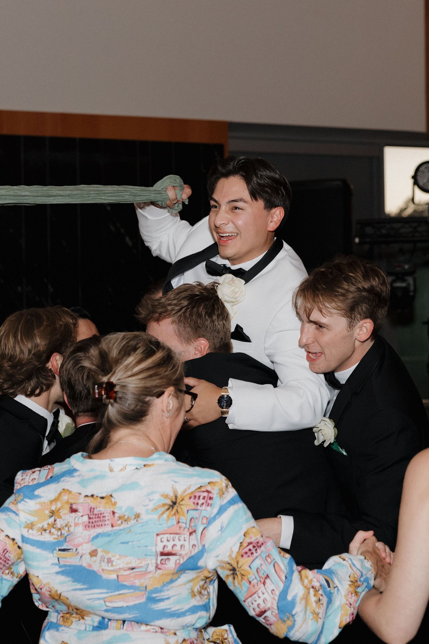 Groomsmen in formal attire dance and celebrate at a wedding reception wearing colorful Hawaiian-style shirts.