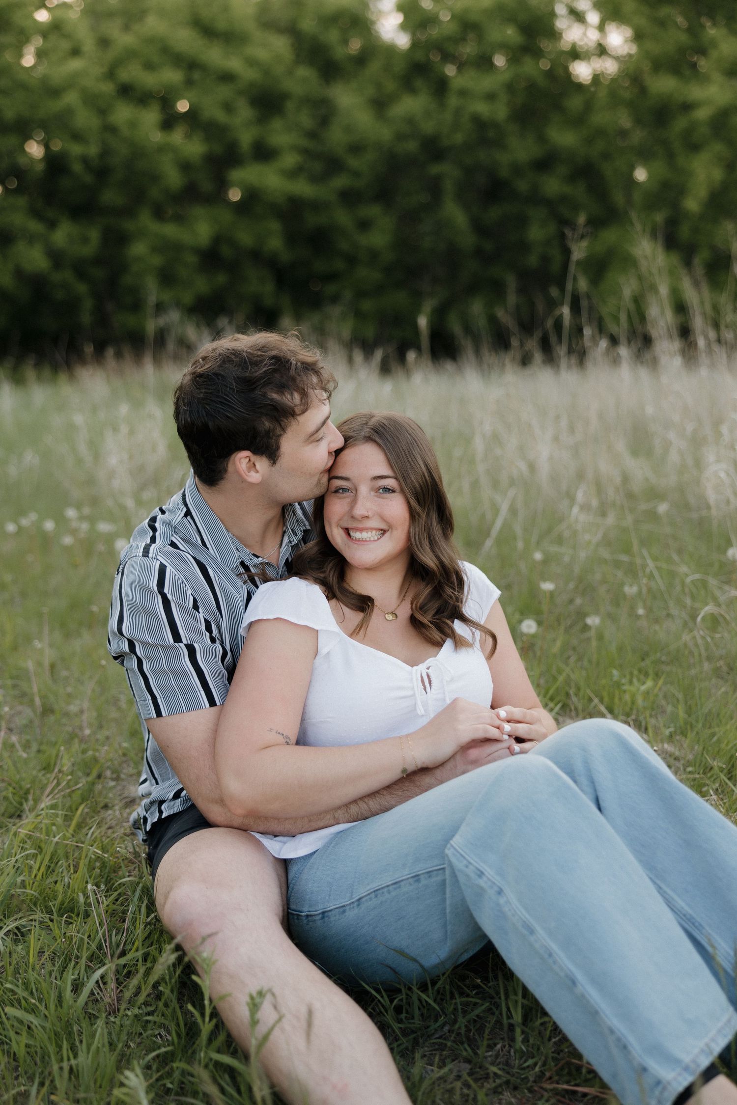 Man in striped shirt kissing woman's cheek as she smiles sitting together in grassy meadow.