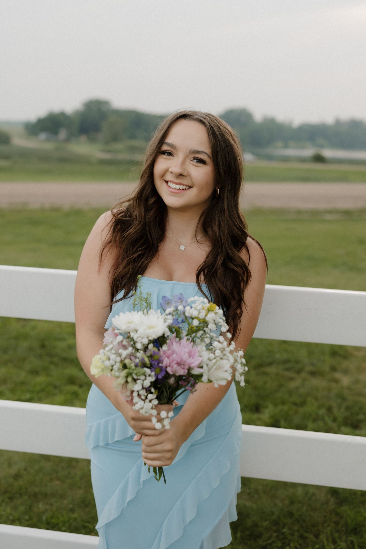 Smiling young woman in light blue ruffle dress holding wildflower bouquet by white fence in green field.