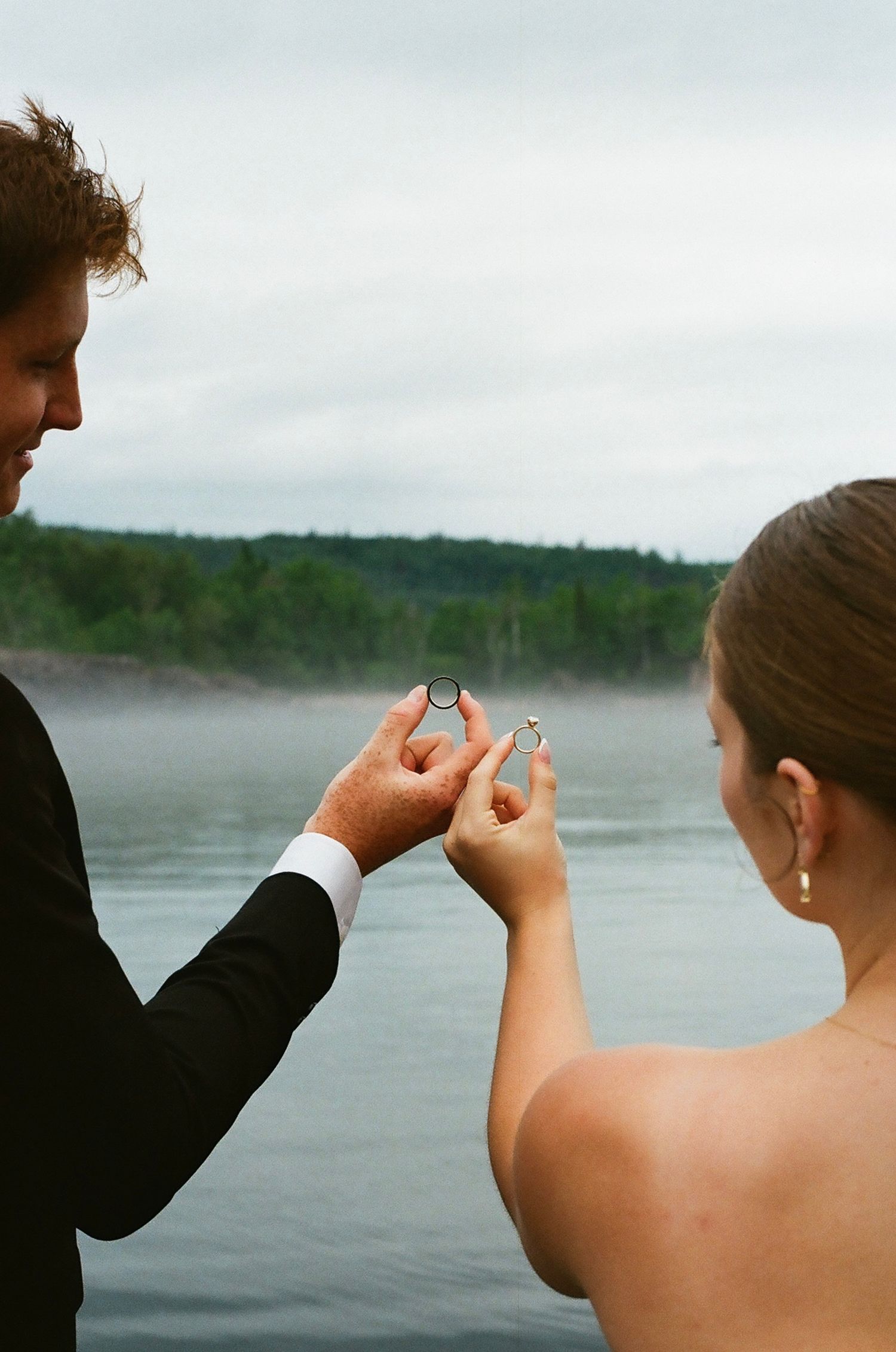 Bride and groom exchanging wedding rings by a misty lake with forested hills in the background.
