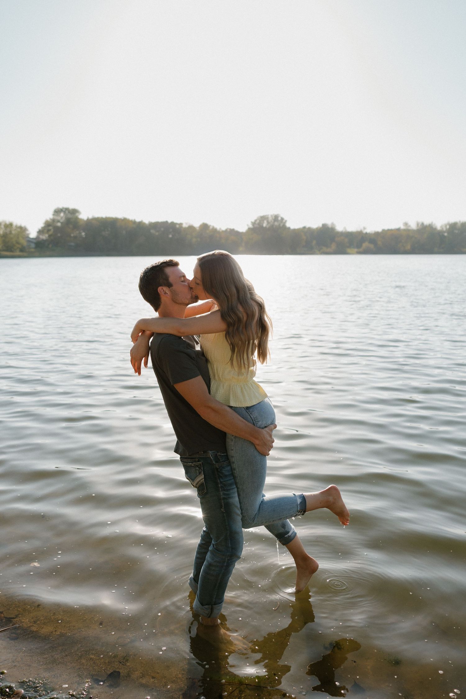 Man lifting woman for a kiss while standing in a sunlit lake shoreline.