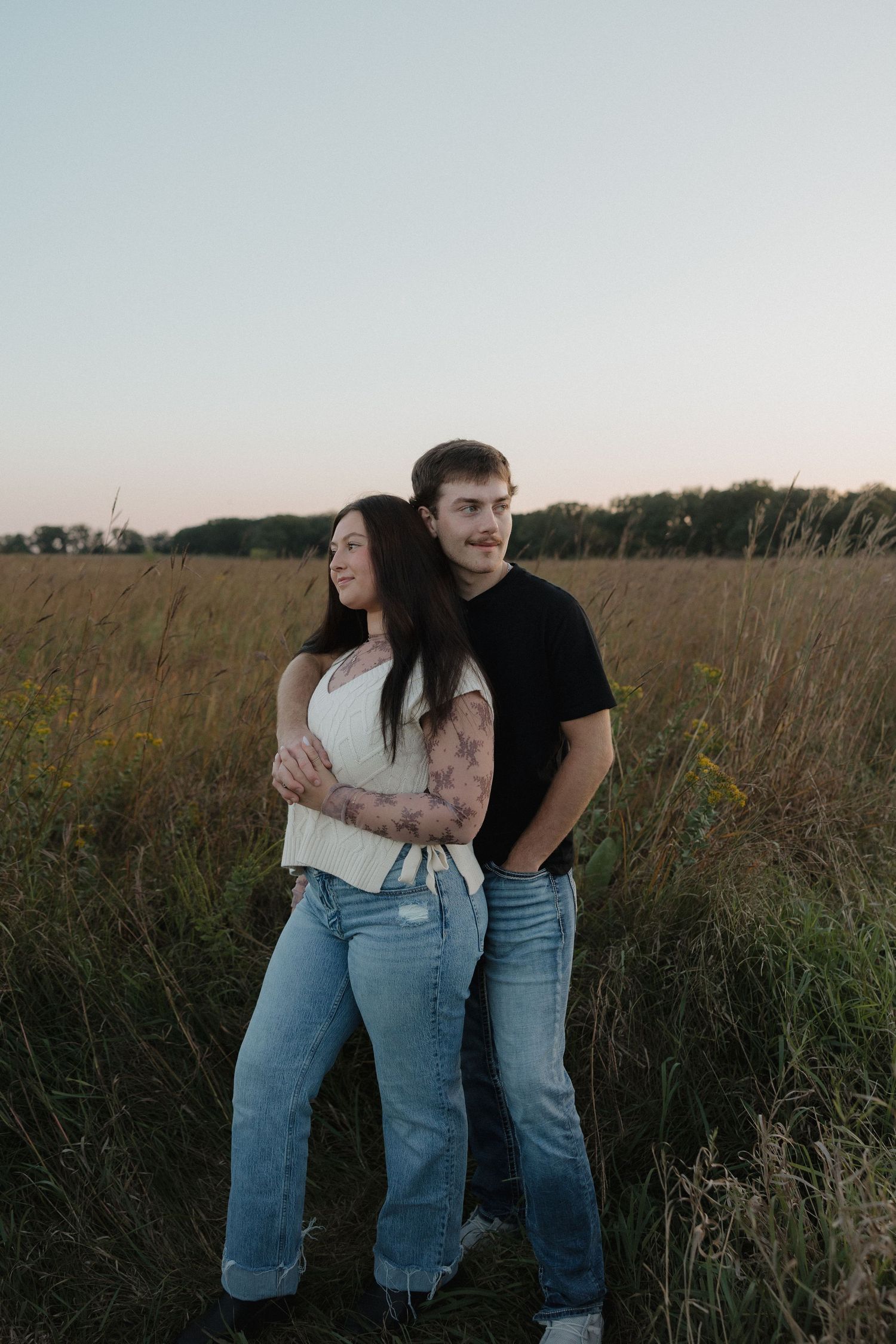 Couple standing in a golden field at sunset, man embracing woman from behind.