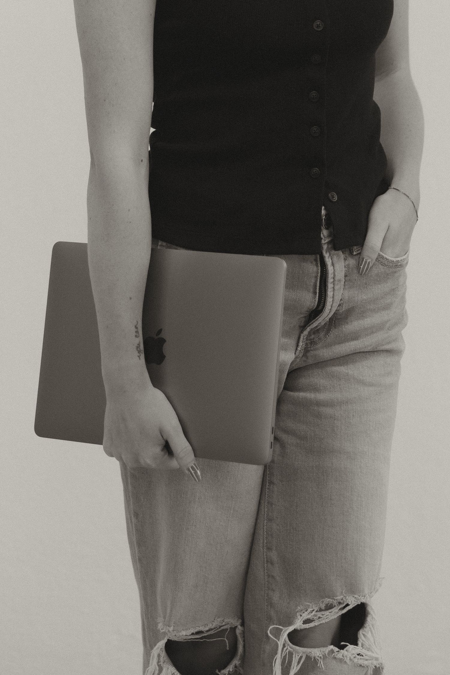 Black and white close-up of woman in ripped jeans holding an Apple MacBook laptop.