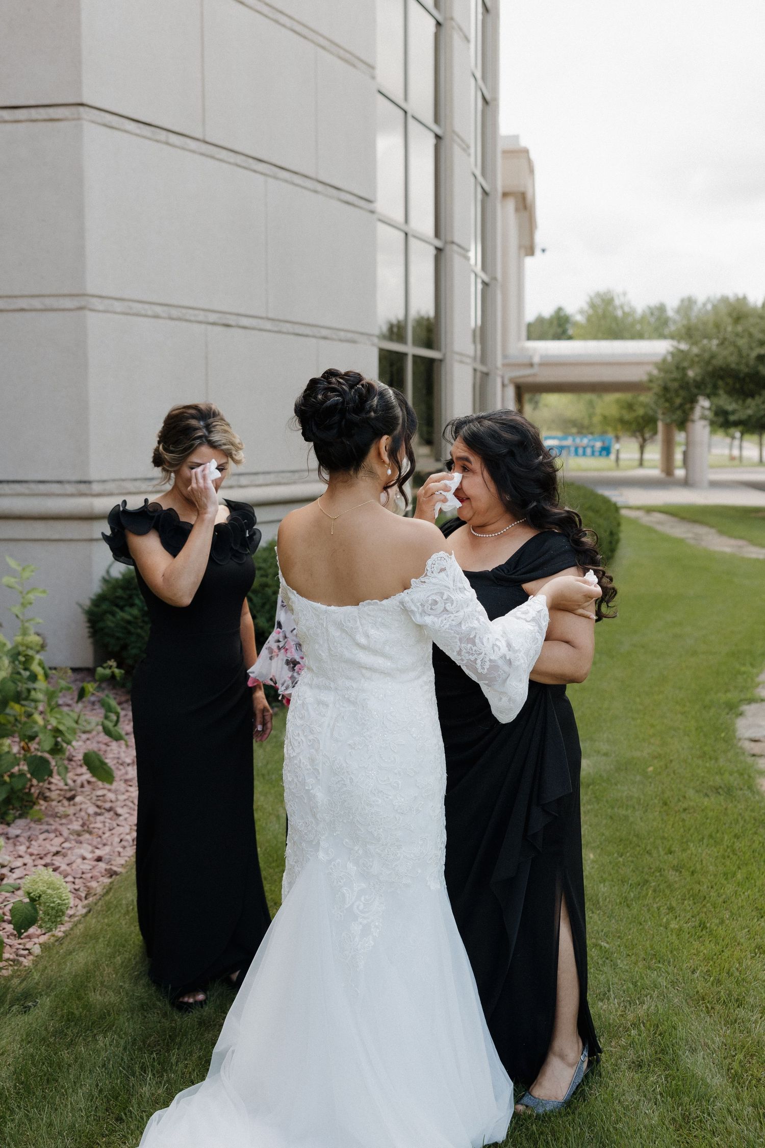 A bride in a lace wedding gown shares an emotional moment with her bridesmaids wearing black dresses.