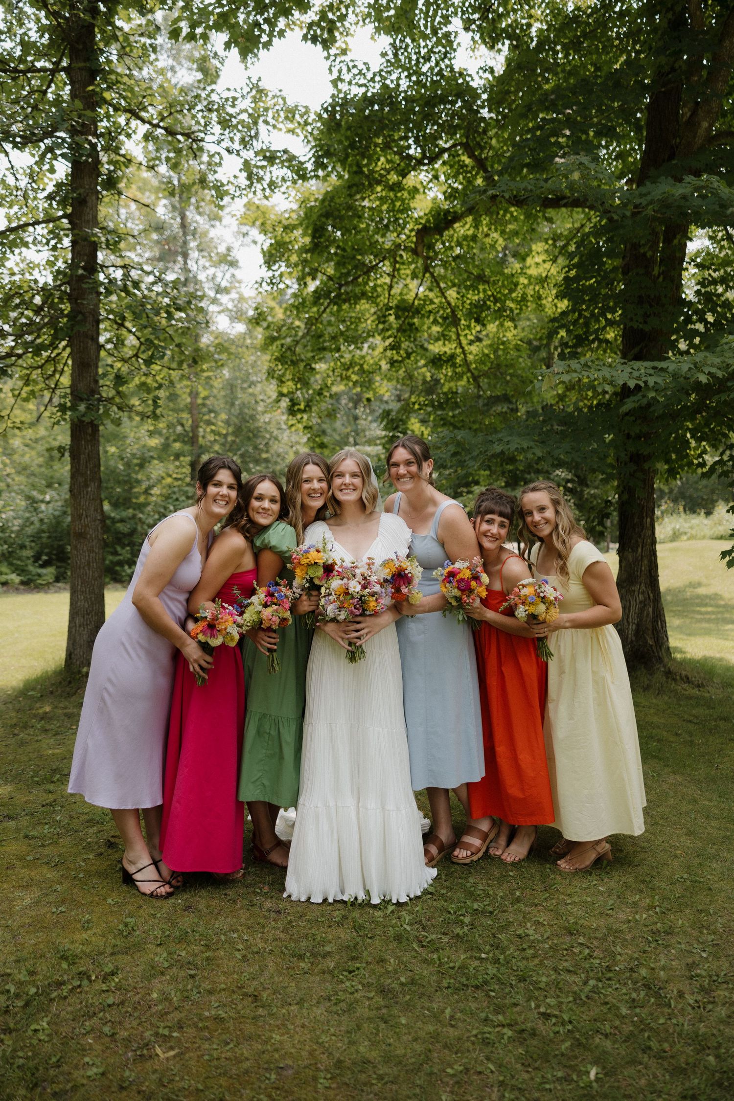 Wedding party poses together outdoors wearing colorful dresses in shades of pink, orange, and sage green.