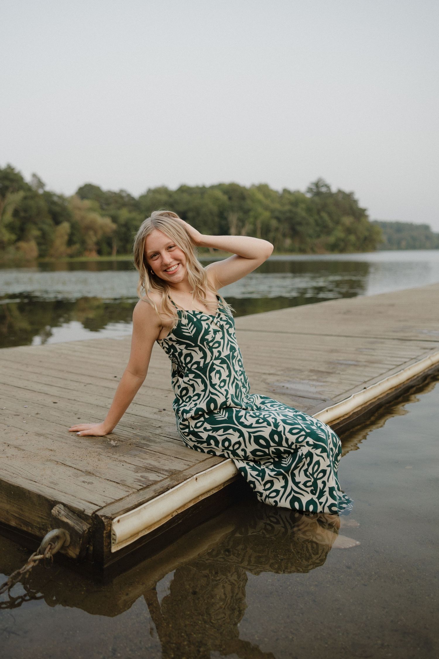 Smiling blonde woman in green patterned dress sitting on wooden dock edge over calm lake with trees behind.