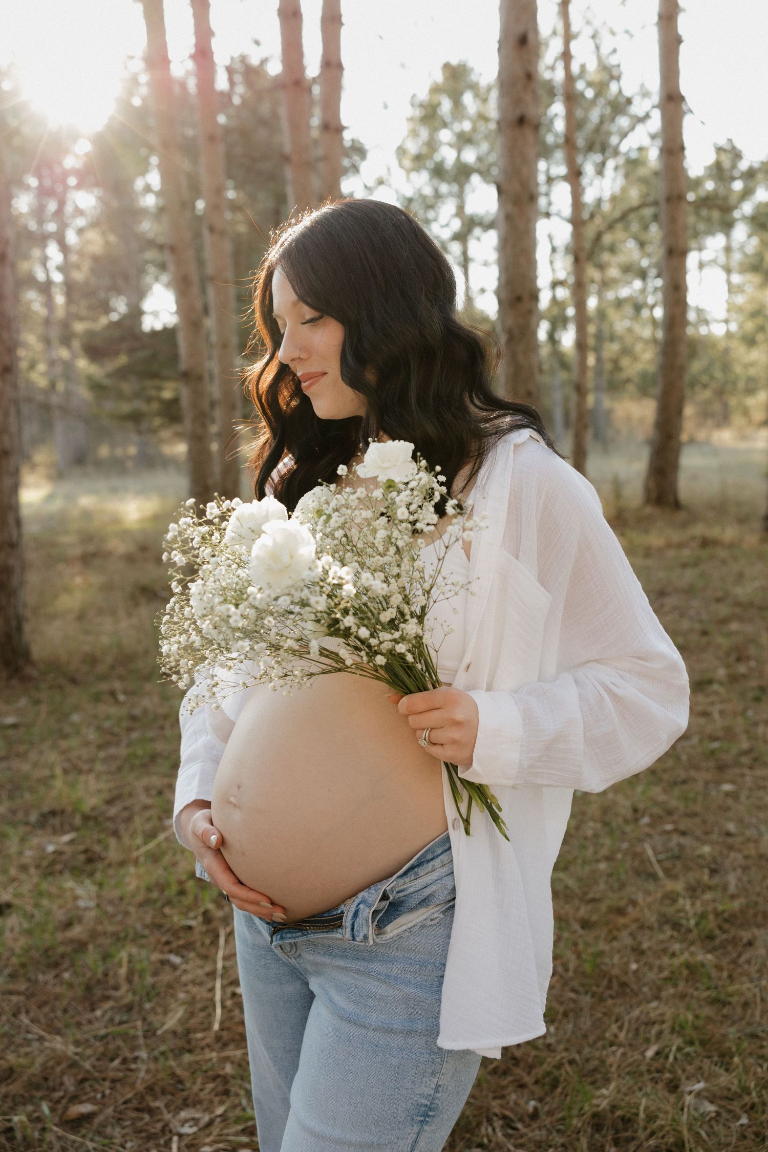 Pregnant woman in white shirt and jeans holding white bouquet, standing in sunlit pine forest smiling.