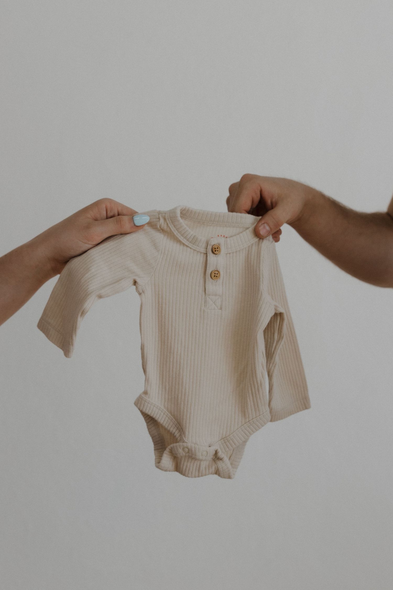 Two hands holding a cream ribbed long-sleeve baby onesie with gold buttons against white background.