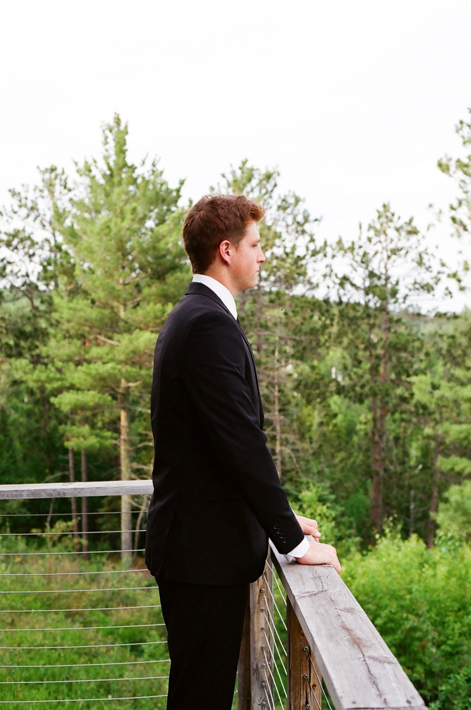 A young man in a black suit stands on a wooden railing deck, gazing at lush green trees in the background.