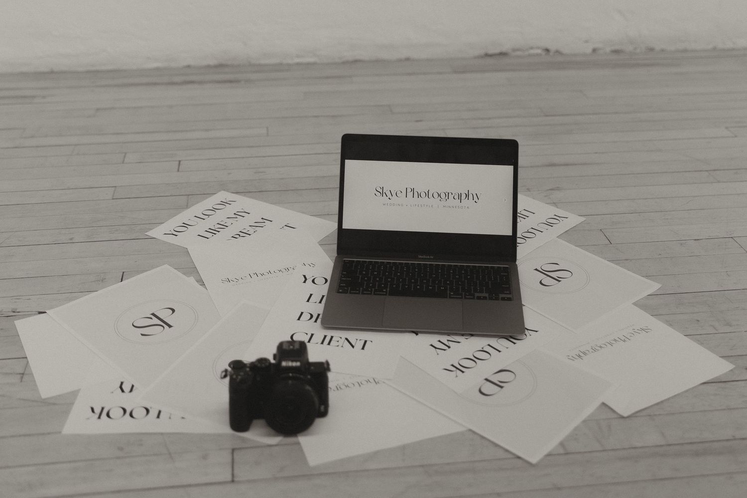 Black and white flat lay of laptop showing photography website, camera, and printed pages.