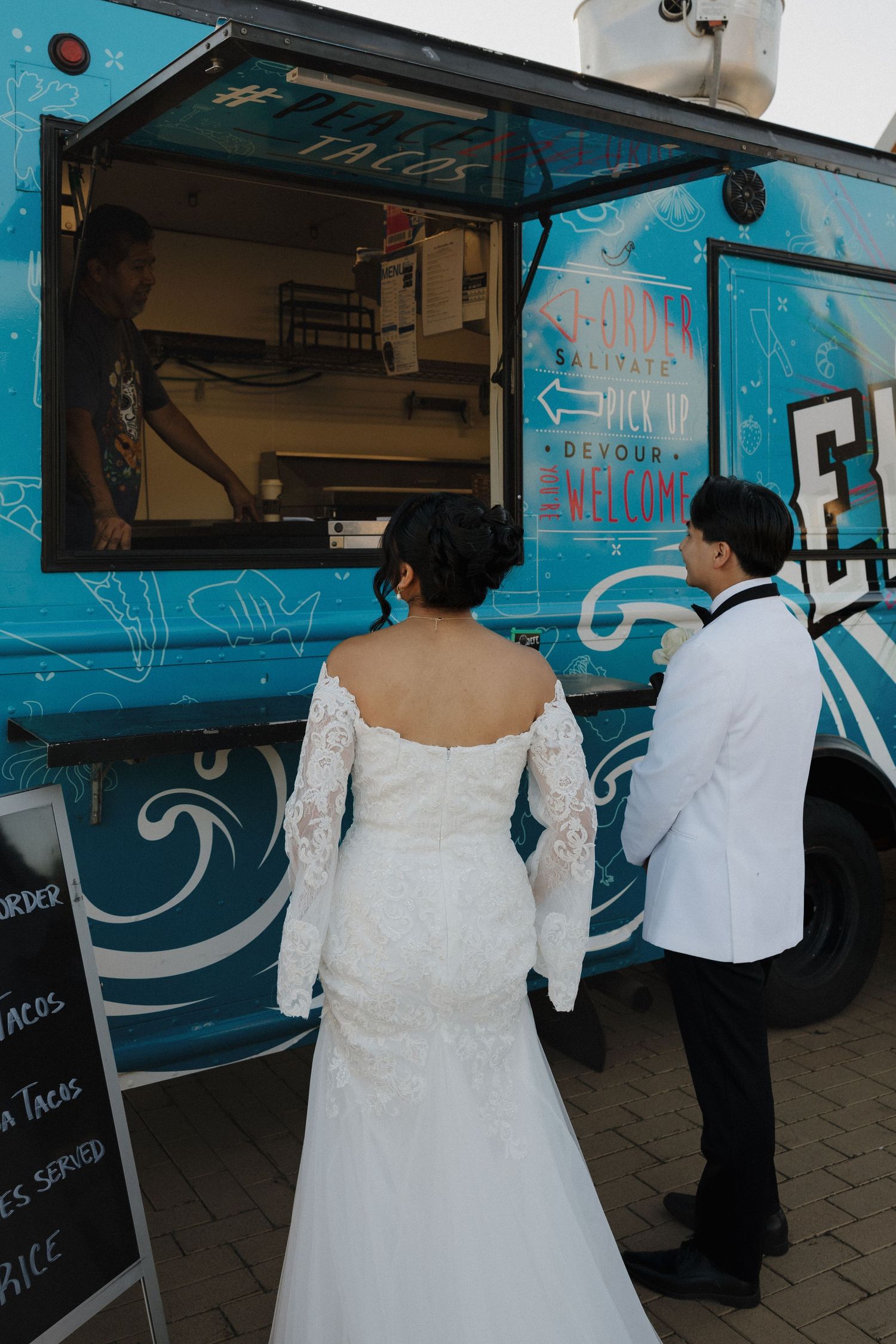 People in formal attire line up at a blue food truck serving refreshments at an outdoor event.