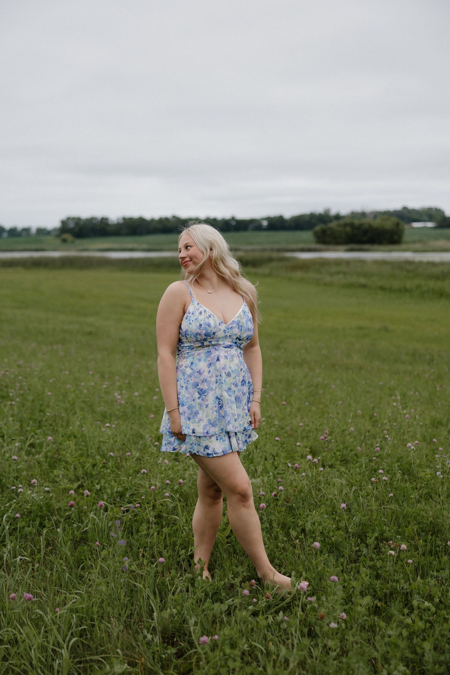 Blonde woman in a blue floral sundress walking barefoot through a green meadow with wildflowers.