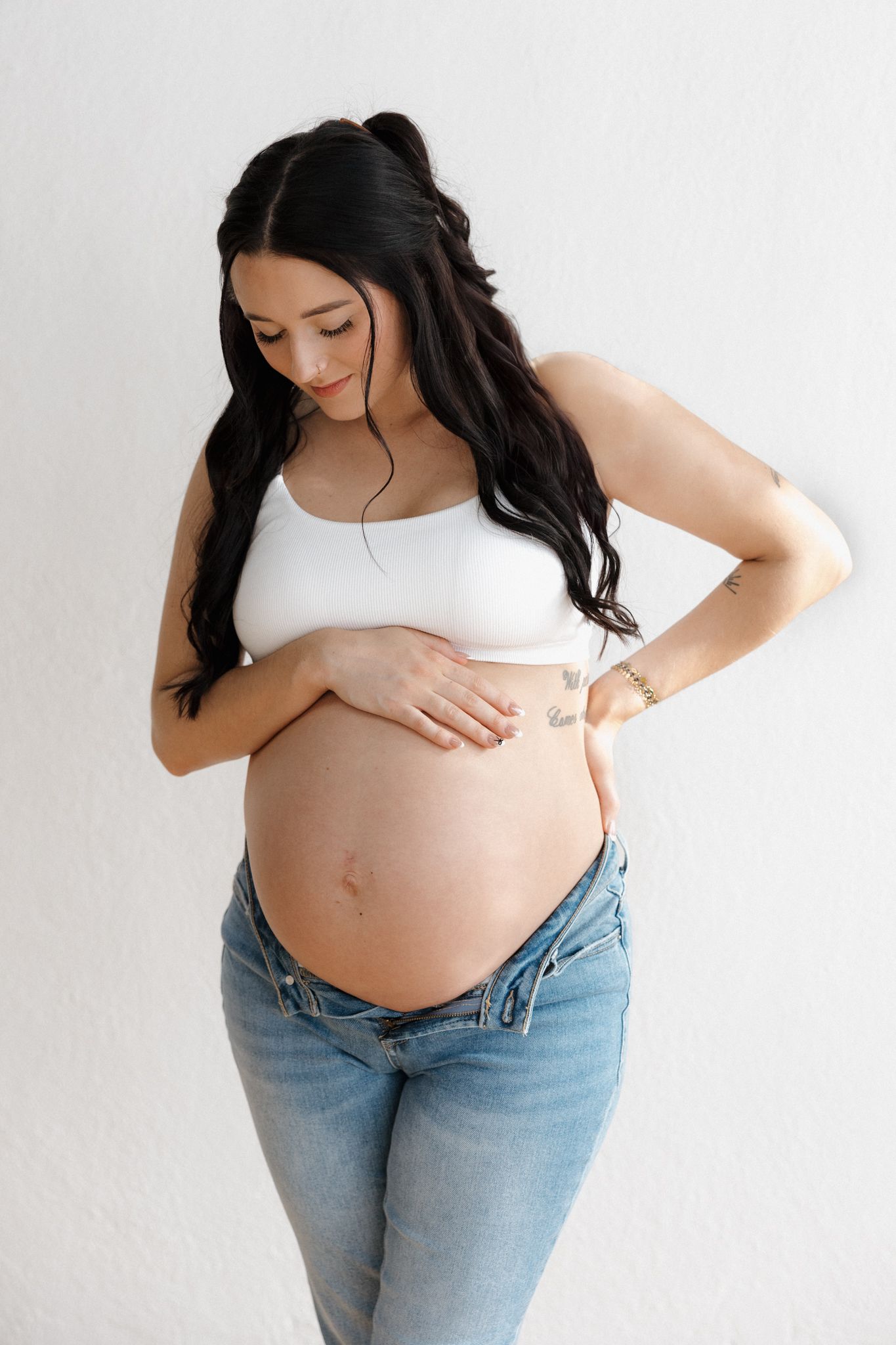 Pregnant woman in white crop top and jeans gently cradling bare baby bump against white wall.