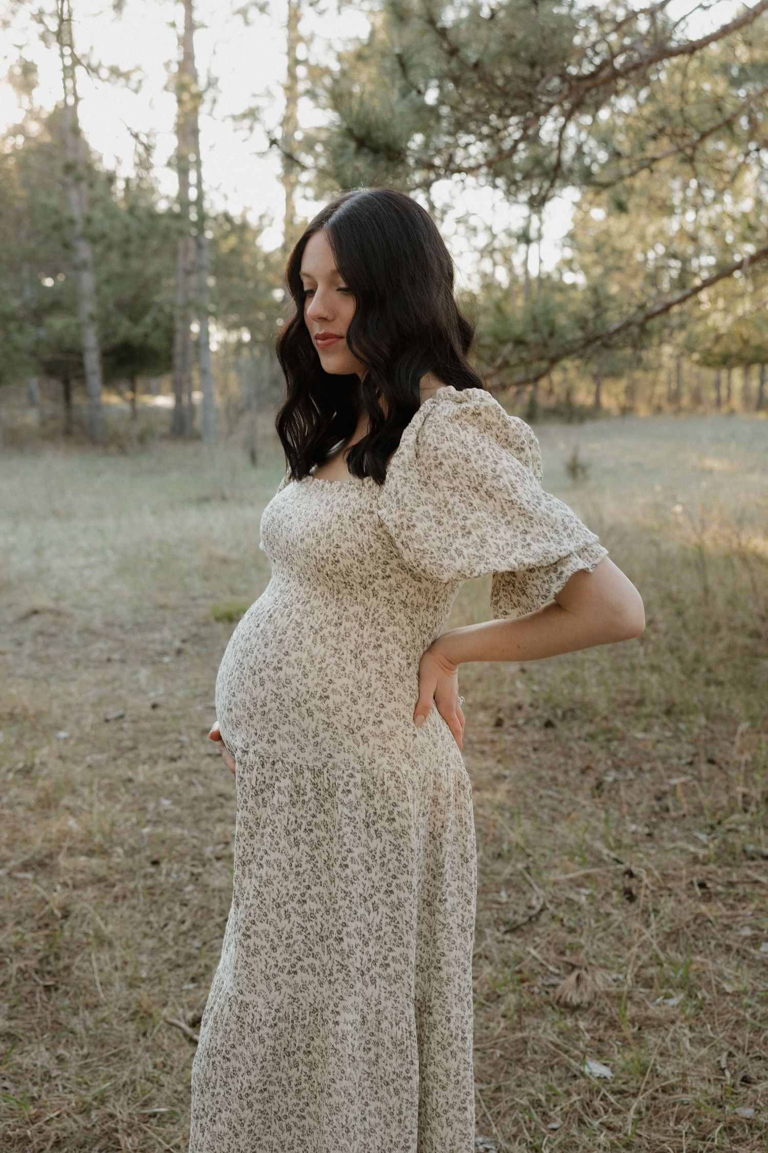Pregnant woman in floral midi dress cradling baby bump in a natural outdoor setting.