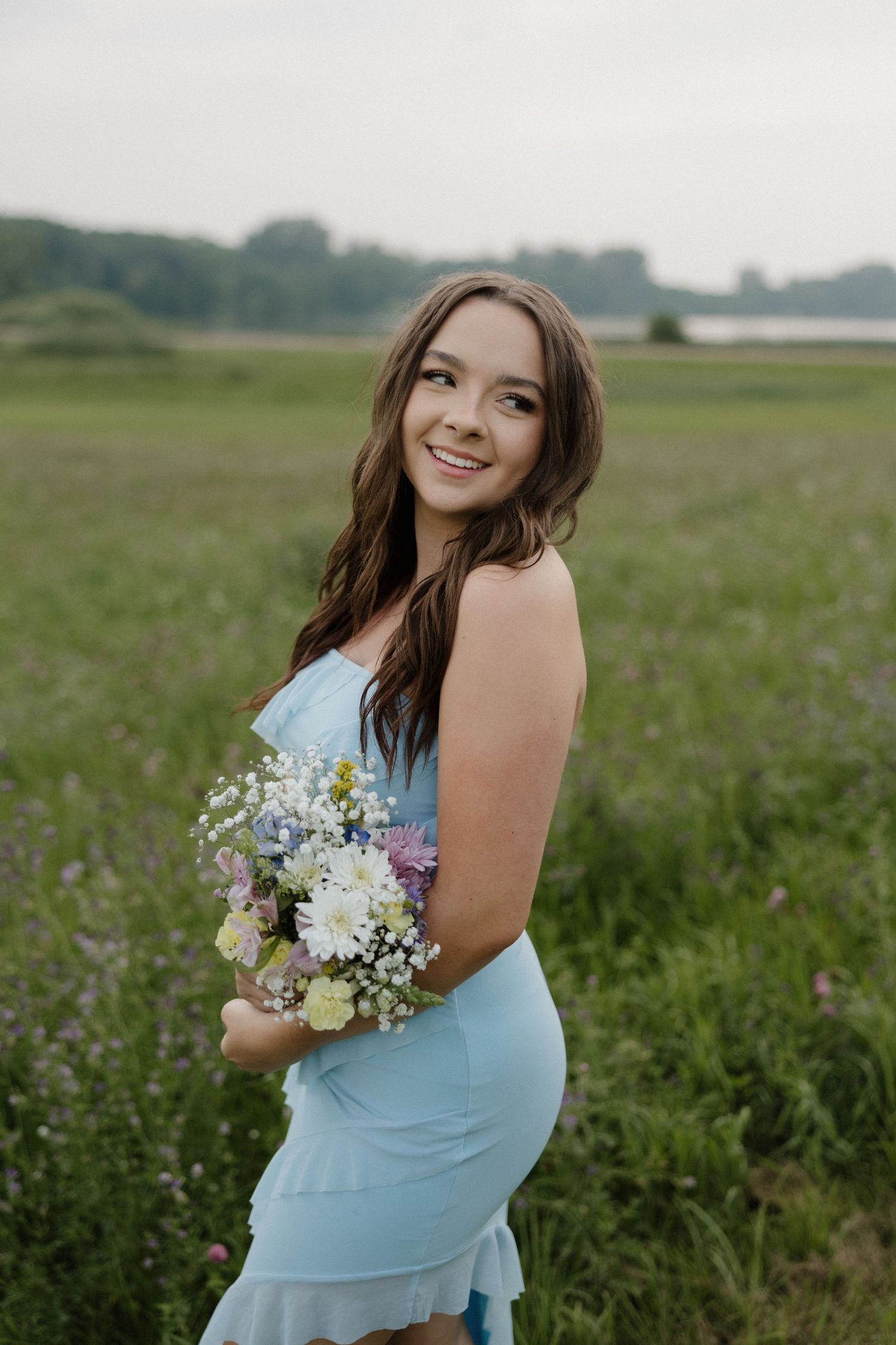 Happy young woman in light blue dress holding wildflower bouquet while standing in lush green meadow.