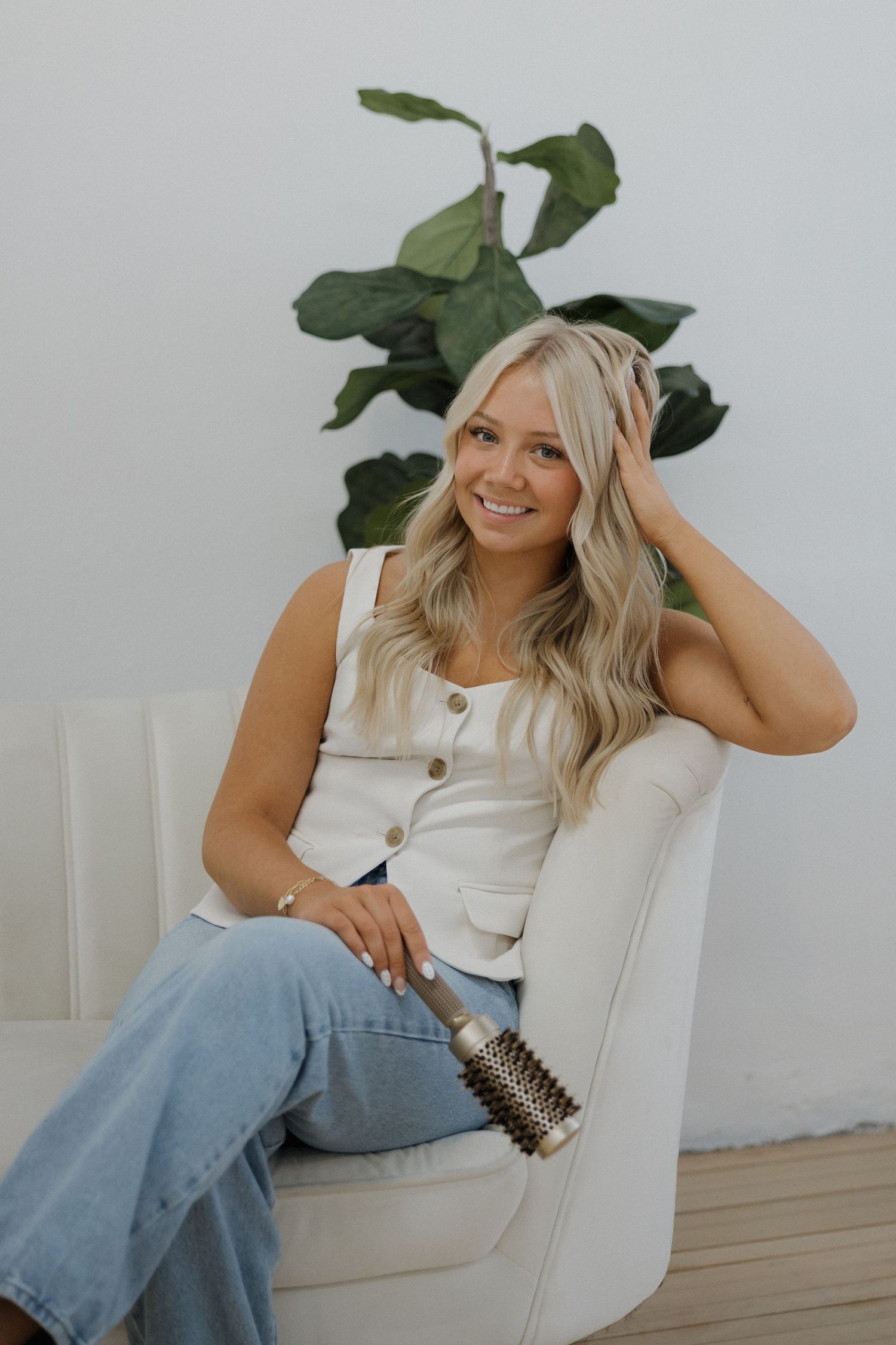 Blonde woman sitting in white chair holding a round brush with a fiddle leaf fig plant.
