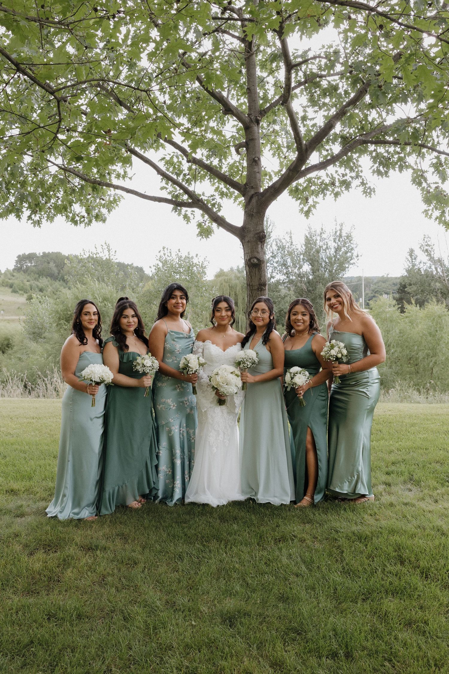 Bridal party in mint and sage dresses poses on grassy hill under tree.