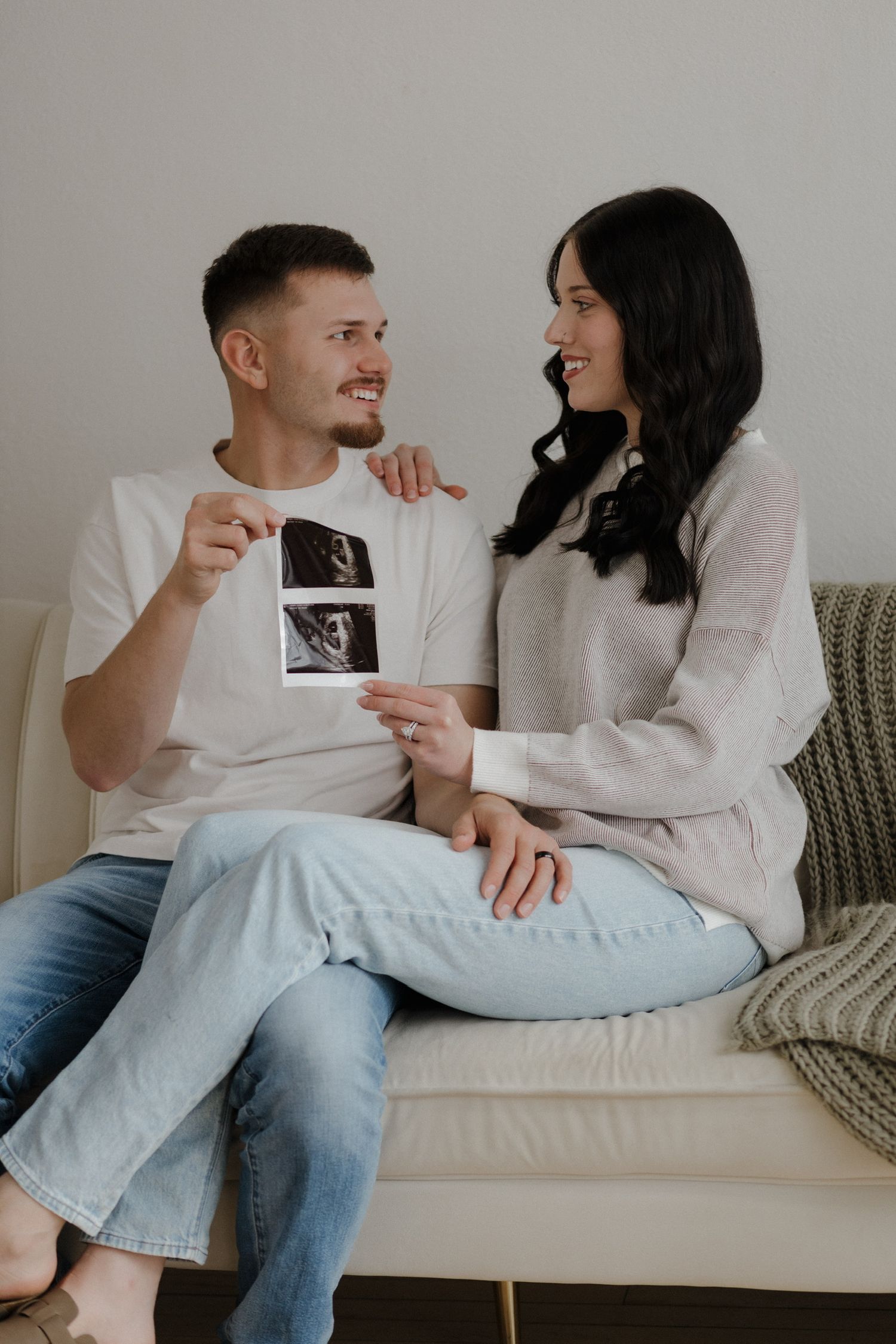 Happy couple sitting on couch smiling at each other while holding ultrasound photos.