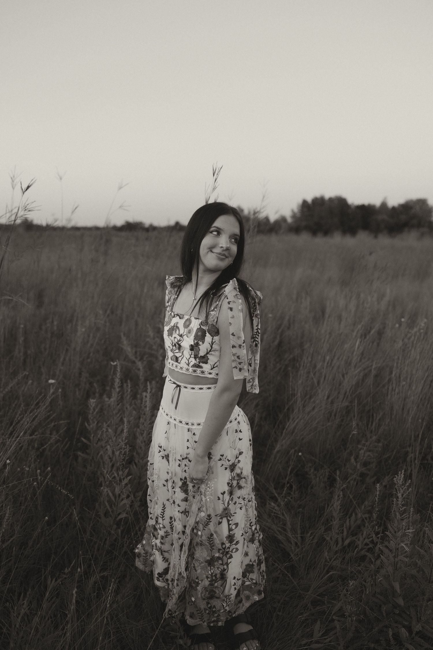 Black and white portrait of dark-haired woman in floral maxi dress standing in open field.