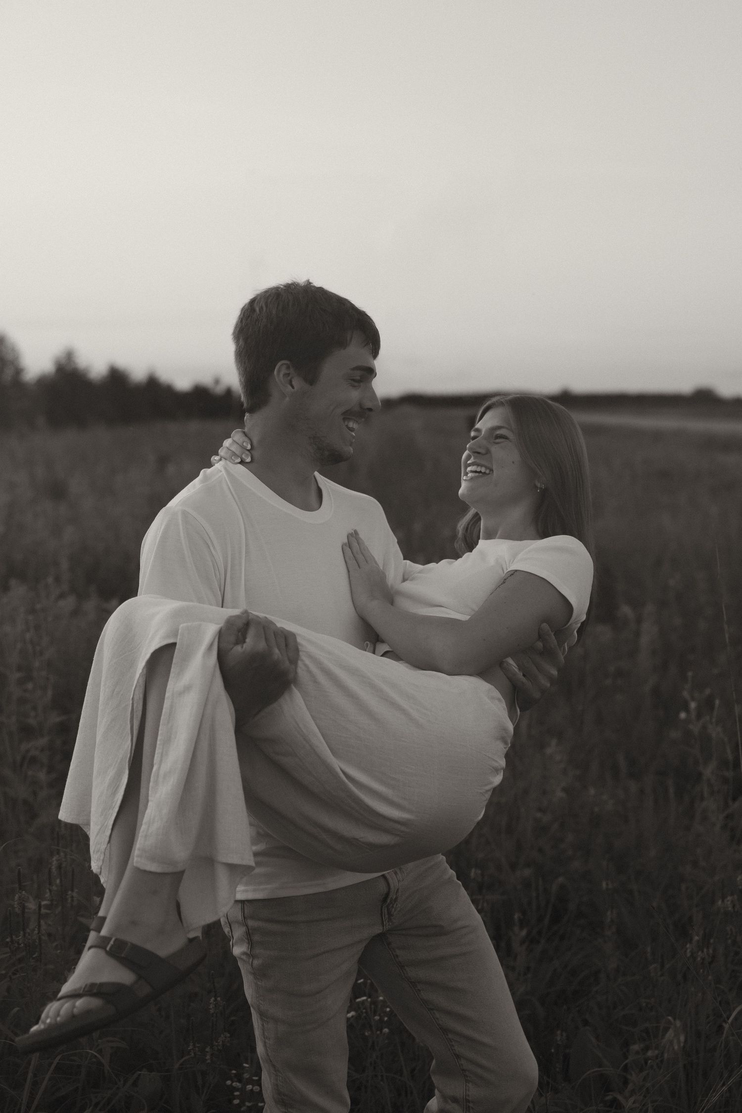 Black and white photo of smiling man carrying laughing woman in his arms in an open field.