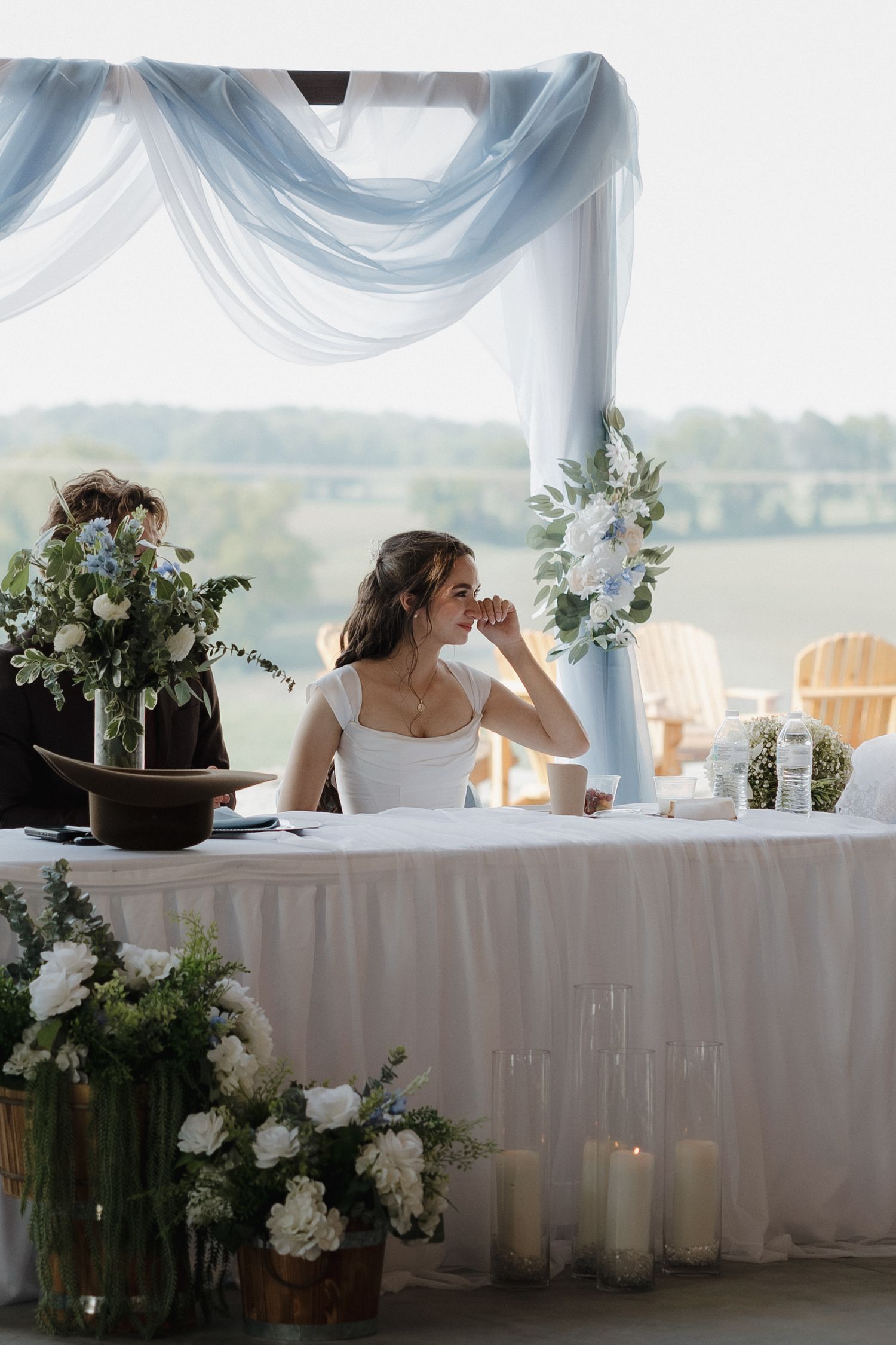 Wedding reception table decorated with white linens and floral arrangements under a draped tent.