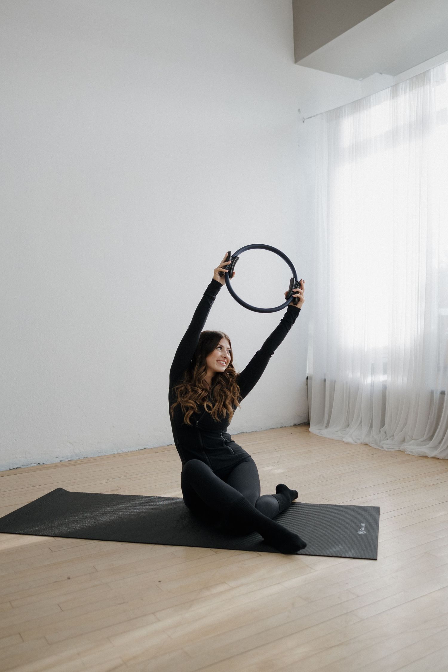 Smiling woman in black workout clothes sitting on yoga mat holding a pilates ring overhead in white studio.