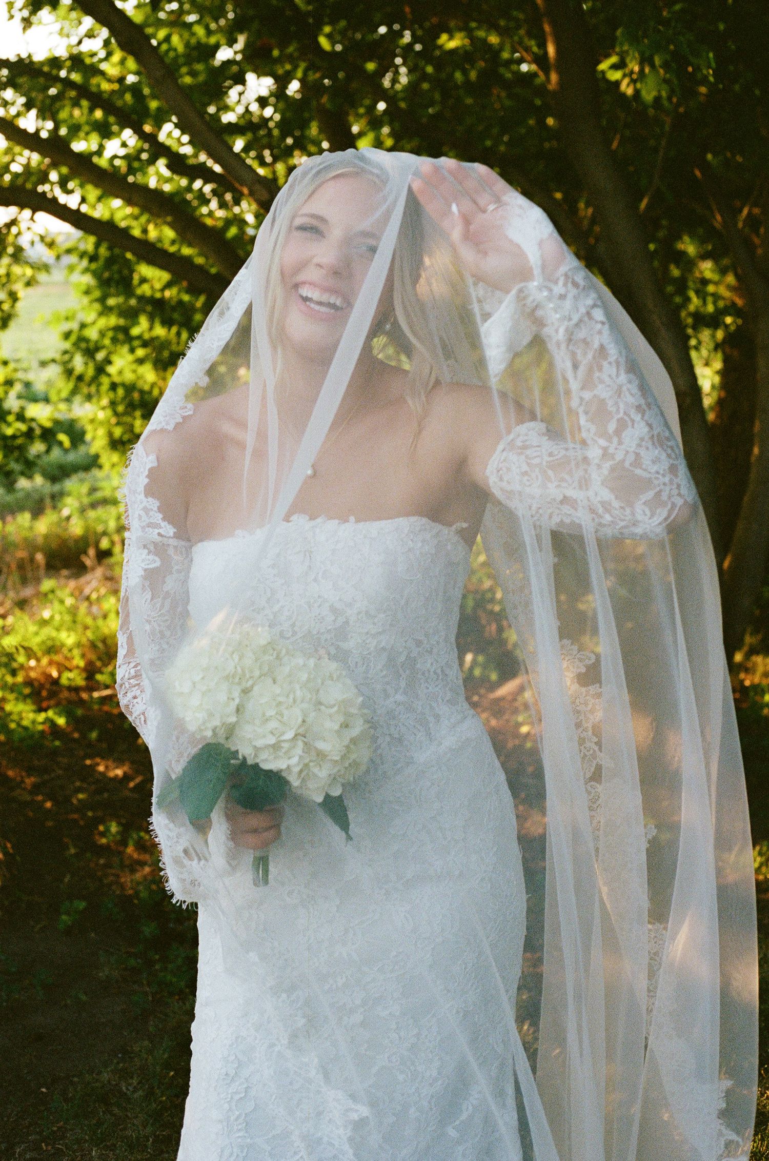 Smiling bride in lace wedding dress and veil holds white hydrangea bouquet outdoors under green trees.