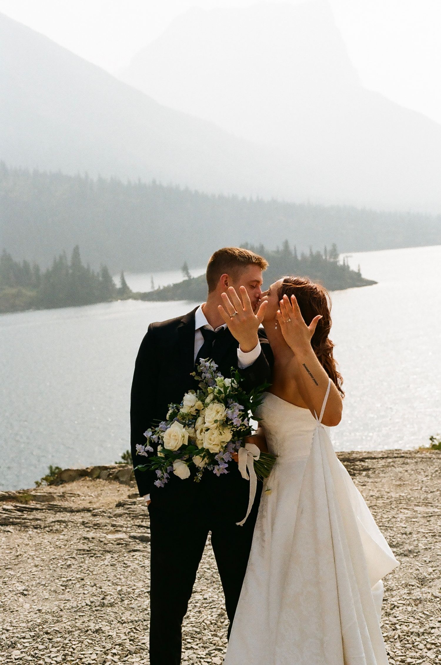 Bride and groom kissing while showing wedding rings, holding bouquet by a mountain lake.