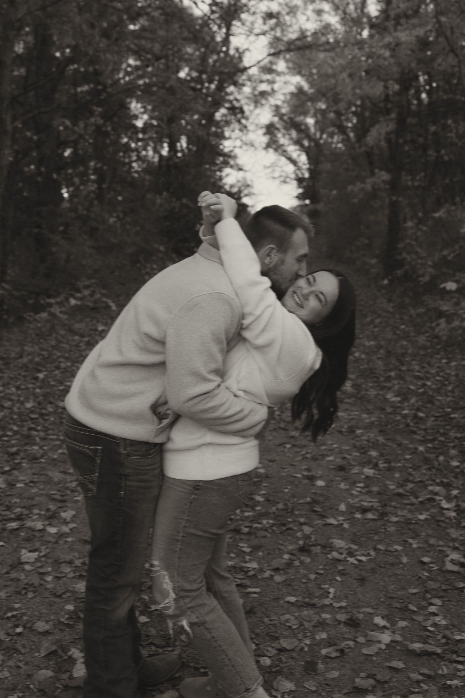 Black and white photo of couple dancing and laughing on a leaf-covered forest path.
