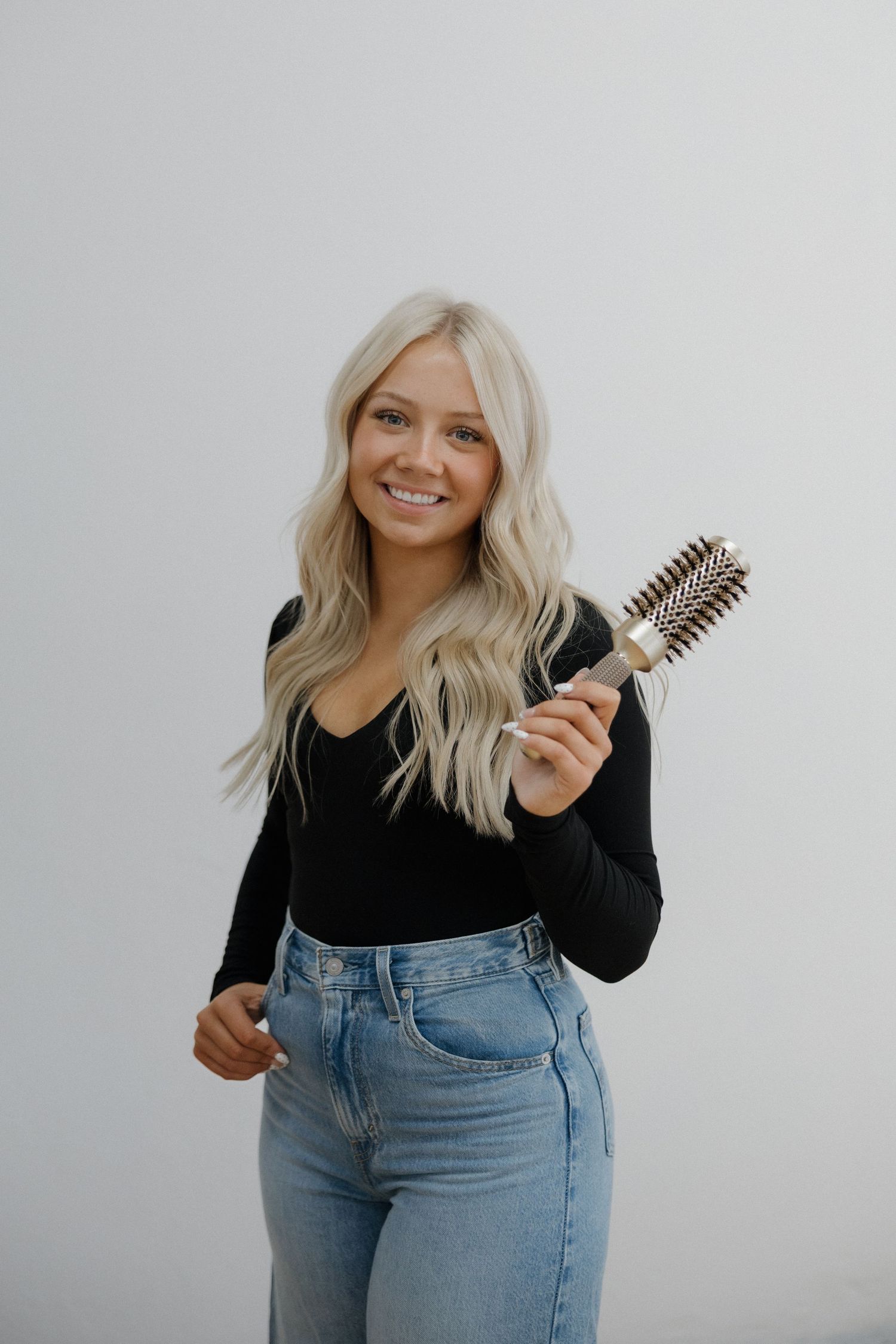Smiling blonde woman in black top and jeans holding a round barrel hair brush.