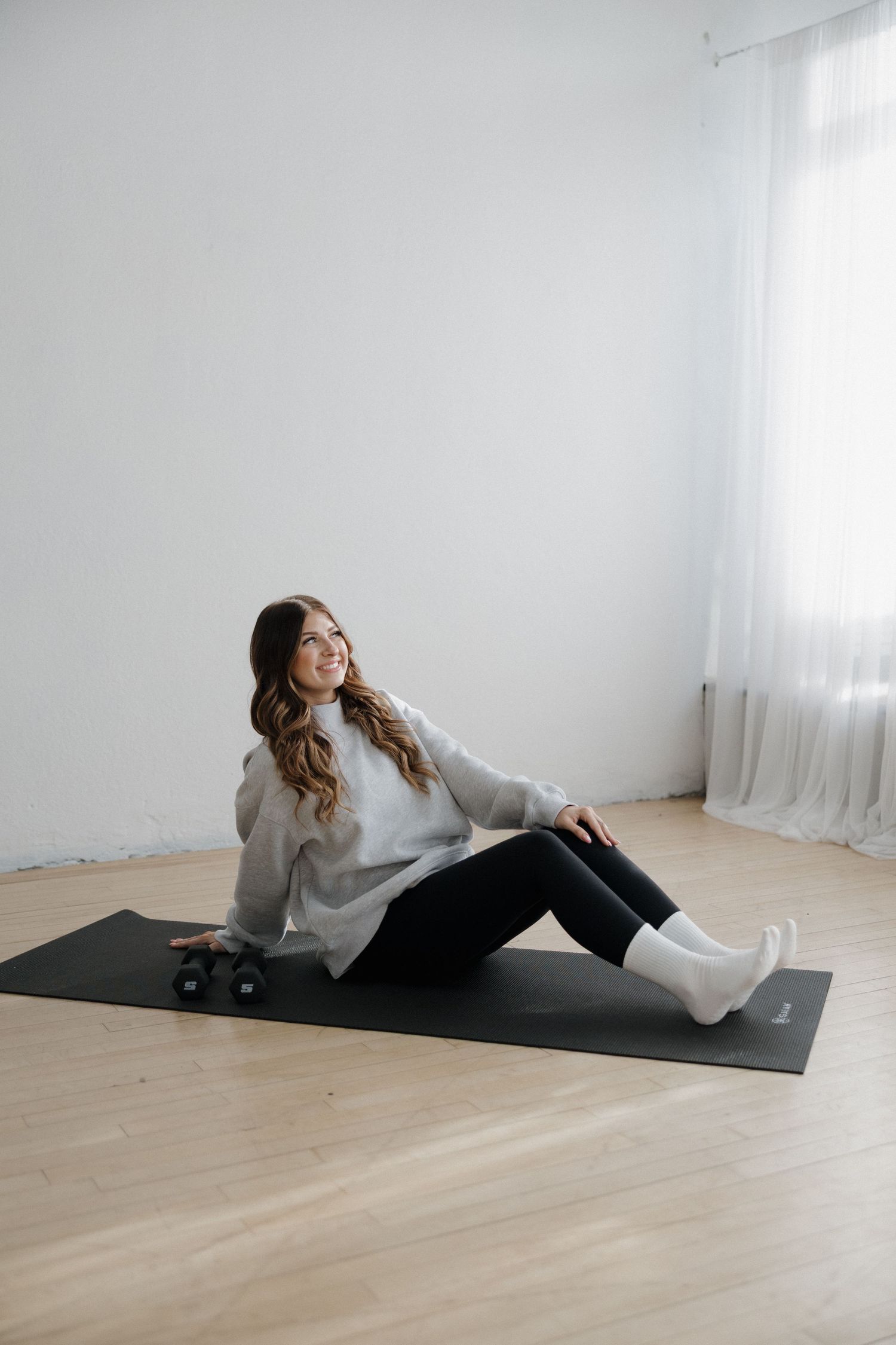 Smiling woman in gray hoodie sitting on black yoga mat with dumbbells in bright white studio space.