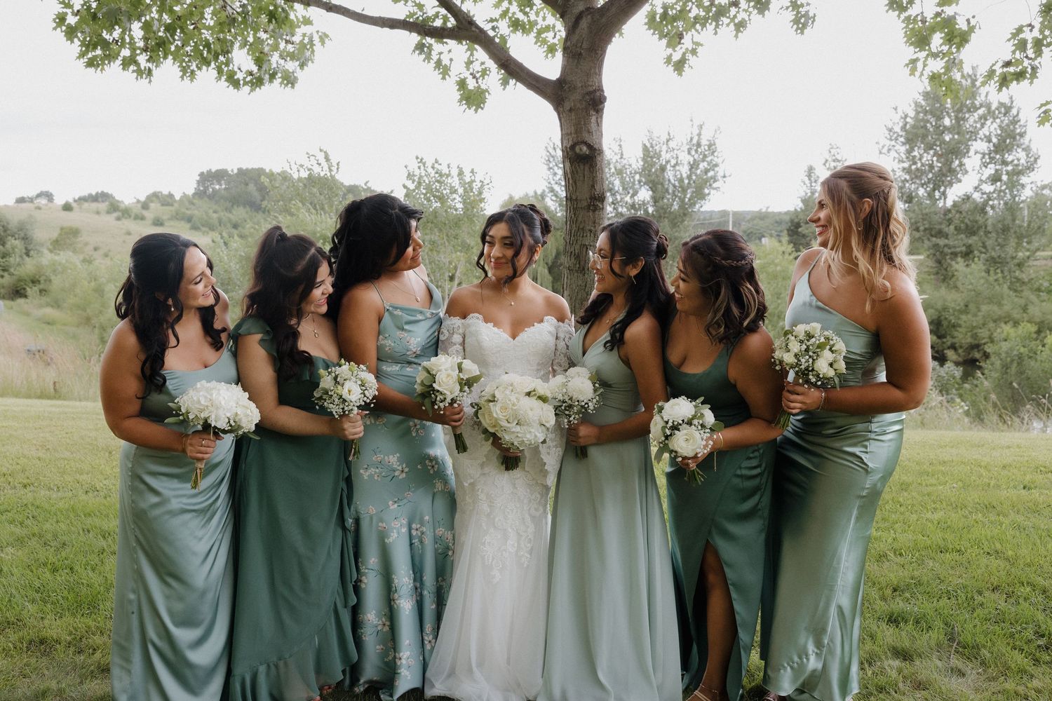 Bridesmaids in sage green dresses pose together outdoors under trees.