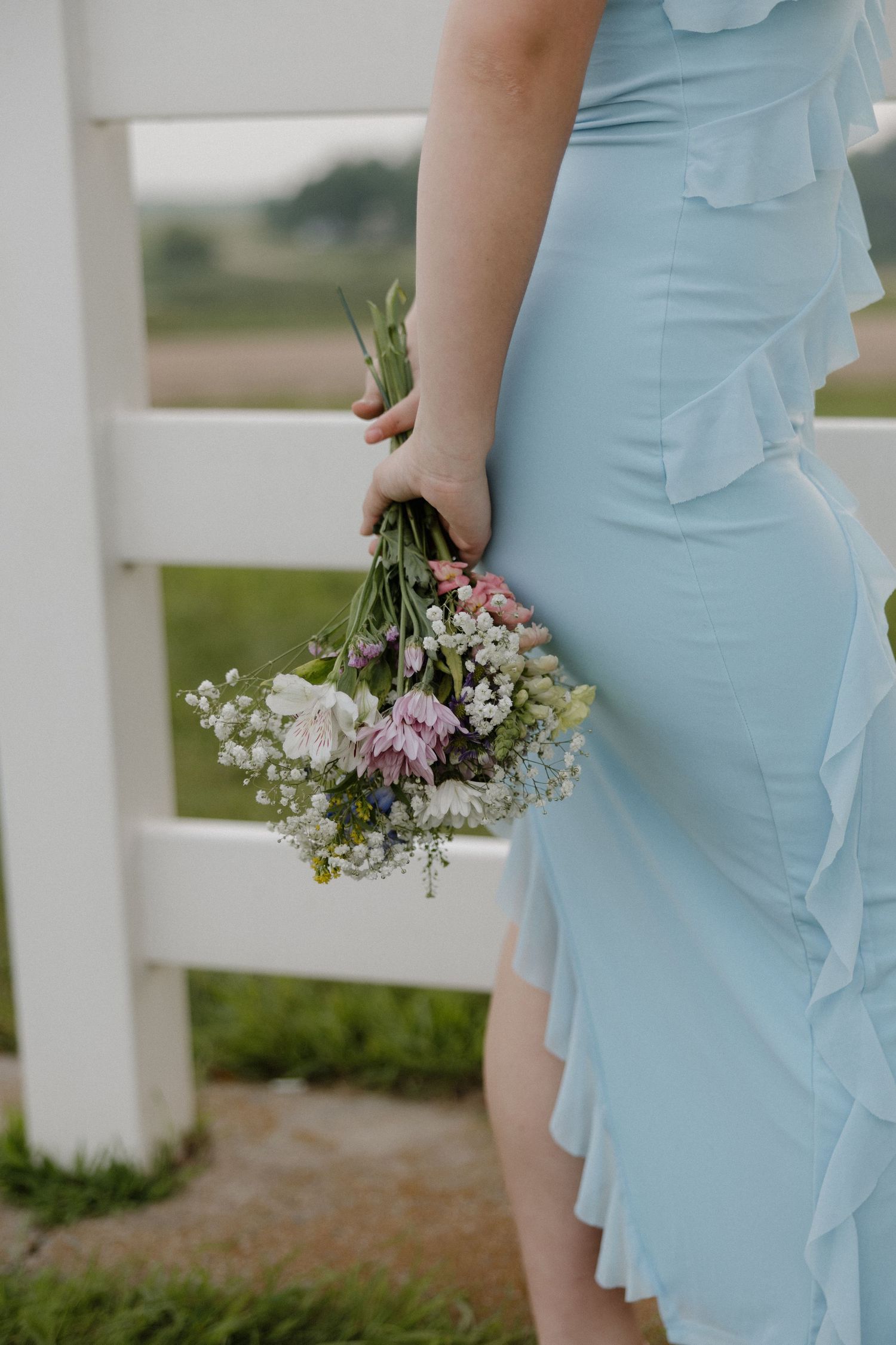 Close-up of woman in light blue ruffle dress holding delicate wildflower bouquet by white wooden fence.